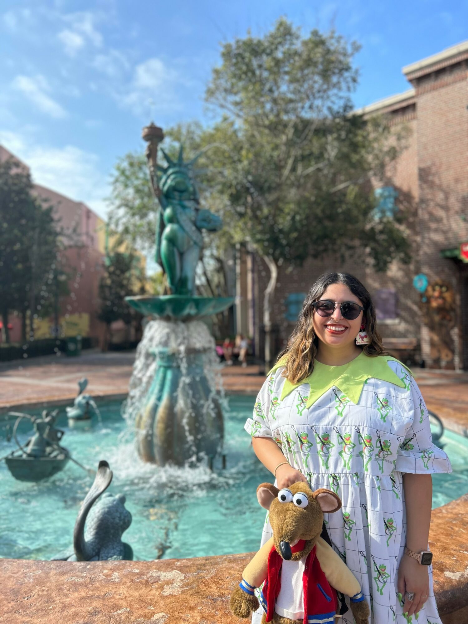 Woman with sunglasses smiling holding a stuffed animal in front of a fountain with a statue, trees, and buildings in the background.