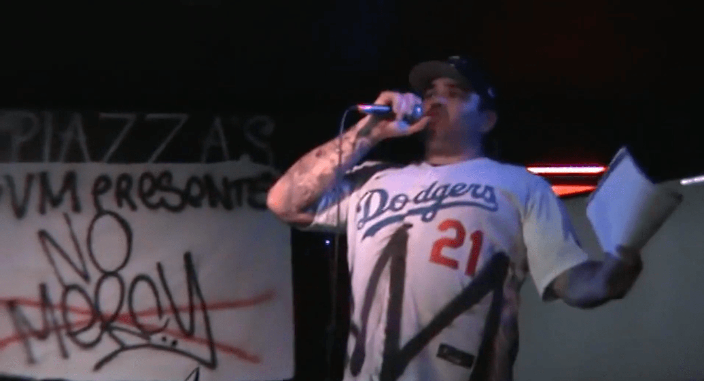 Person holding a microphone and a paper, wearing a Dodgers jersey, on stage with graffiti background.