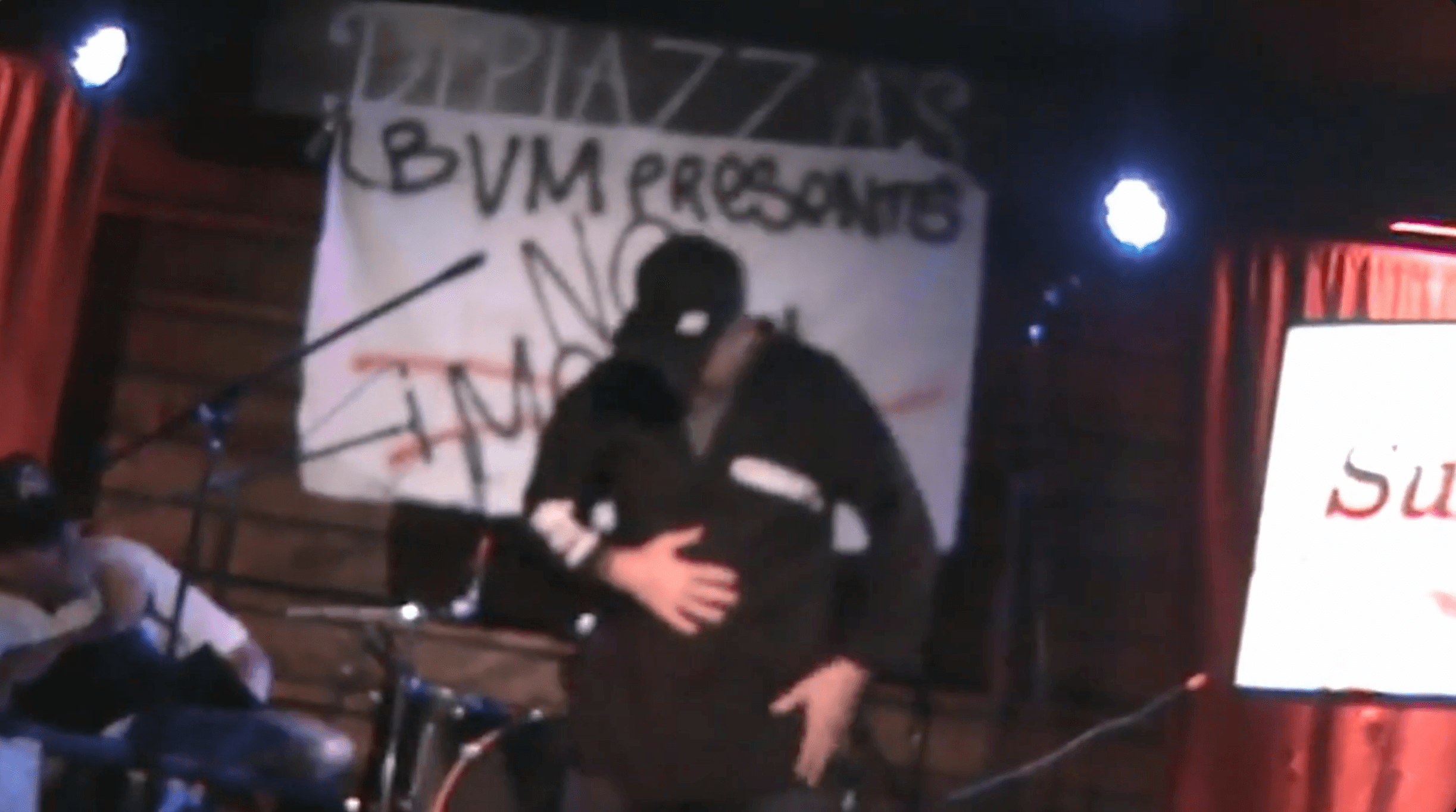 Person on stage with a black hoodie and cap, in front of a banner with text, stage lights visible.