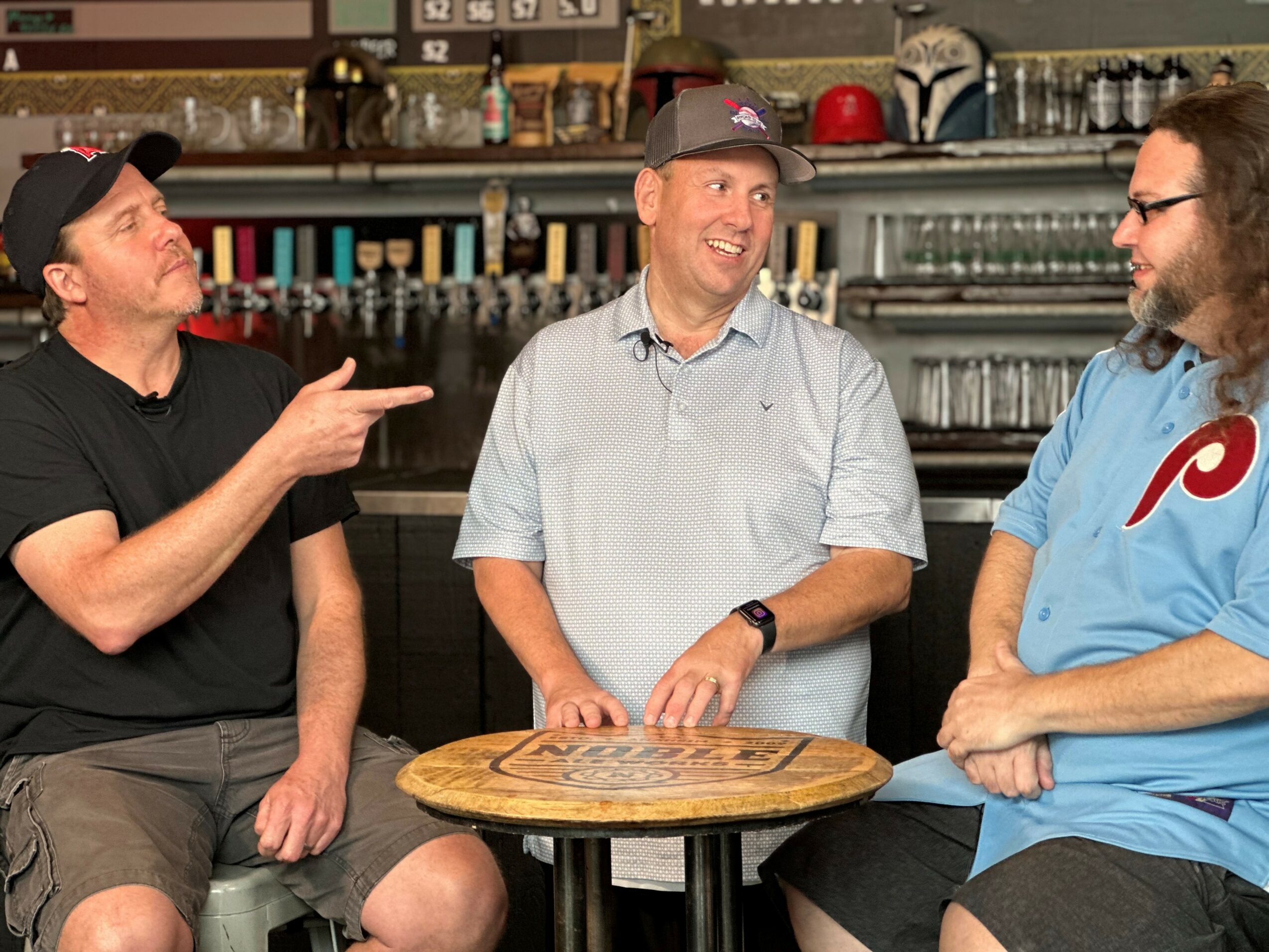 Three men are sitting around a small round table in a bar, engaging in conversation and smiling.