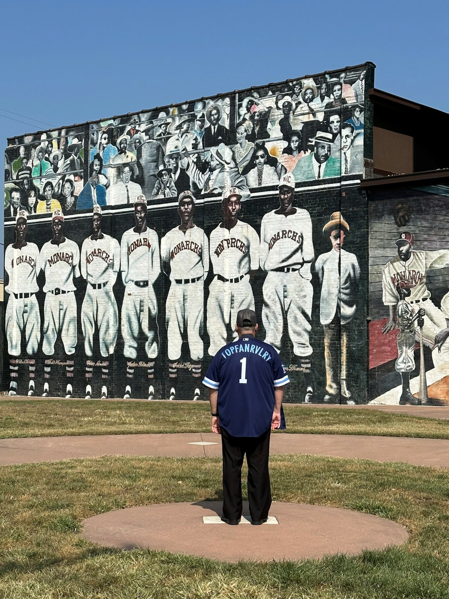 Person wearing a blue jersey stands on a baseball mound facing a large mural of baseball players on a building wall.