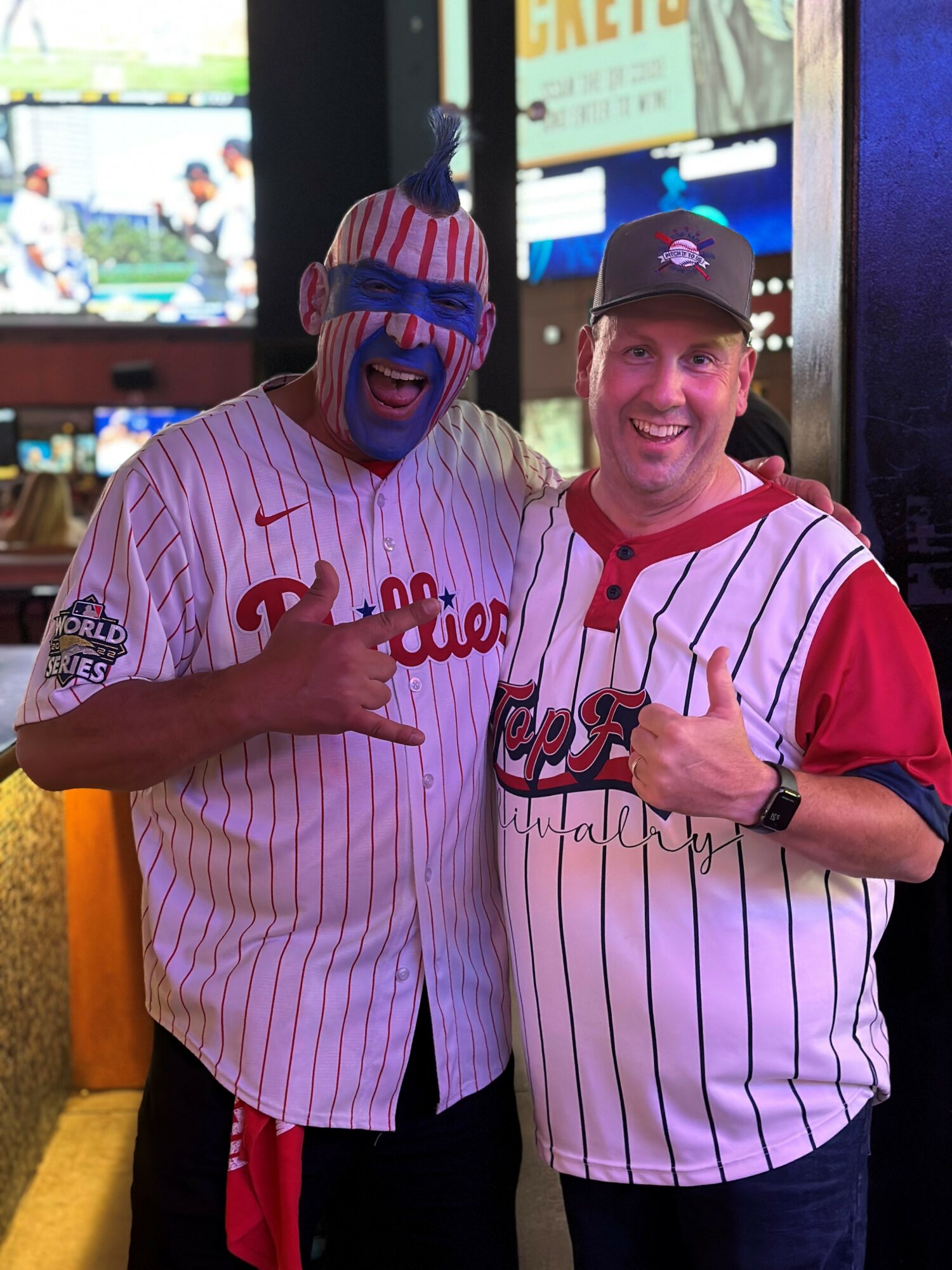 Two people in baseball jerseys posing together, one with face paint and the other wearing a cap, both smiling and making hand gestures.
