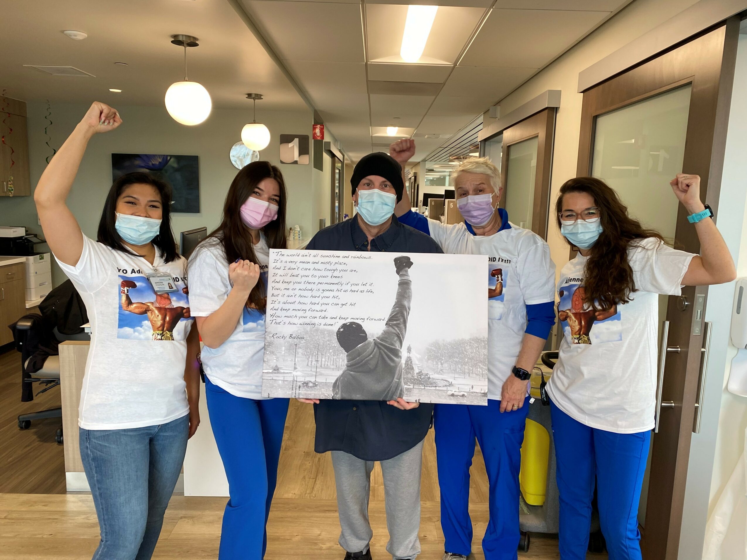 Group of five people wearing masks and matching T-shirts, standing indoors with arms raised in celebration.
