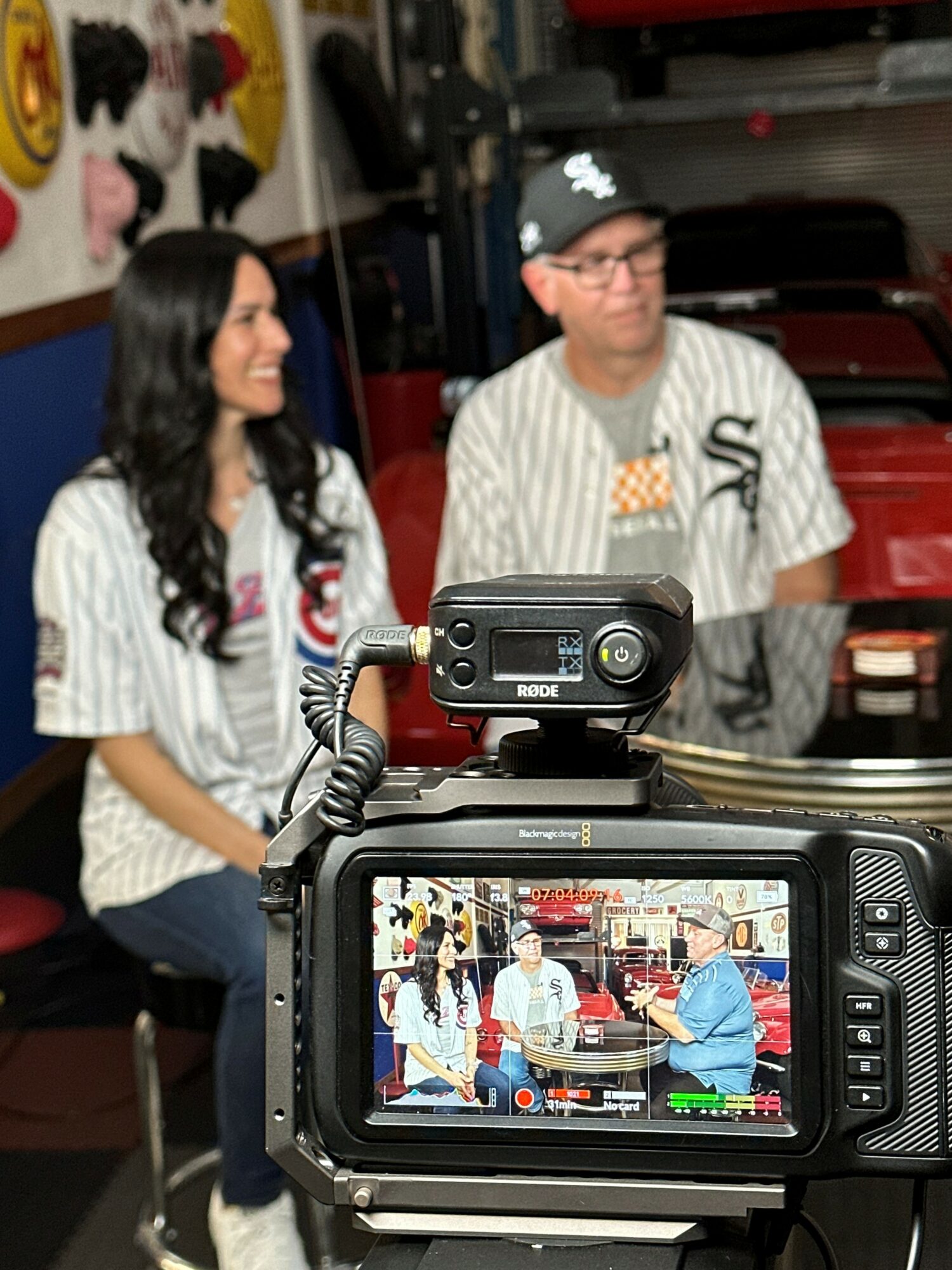 Two people in striped shirts sit in front of a camera, with a camera viewfinder showing them and a red vehicle behind them.