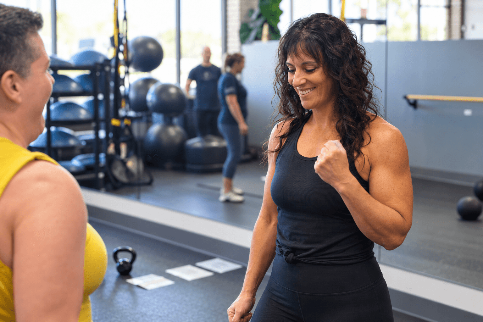 Two women in a gym, one smiling and fist-pumping, with exercise equipment and other people in the background.