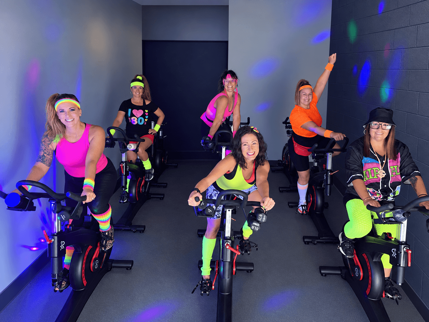 Six women in colorful workout outfits on stationary bikes in a room with dark walls and colorful lights.