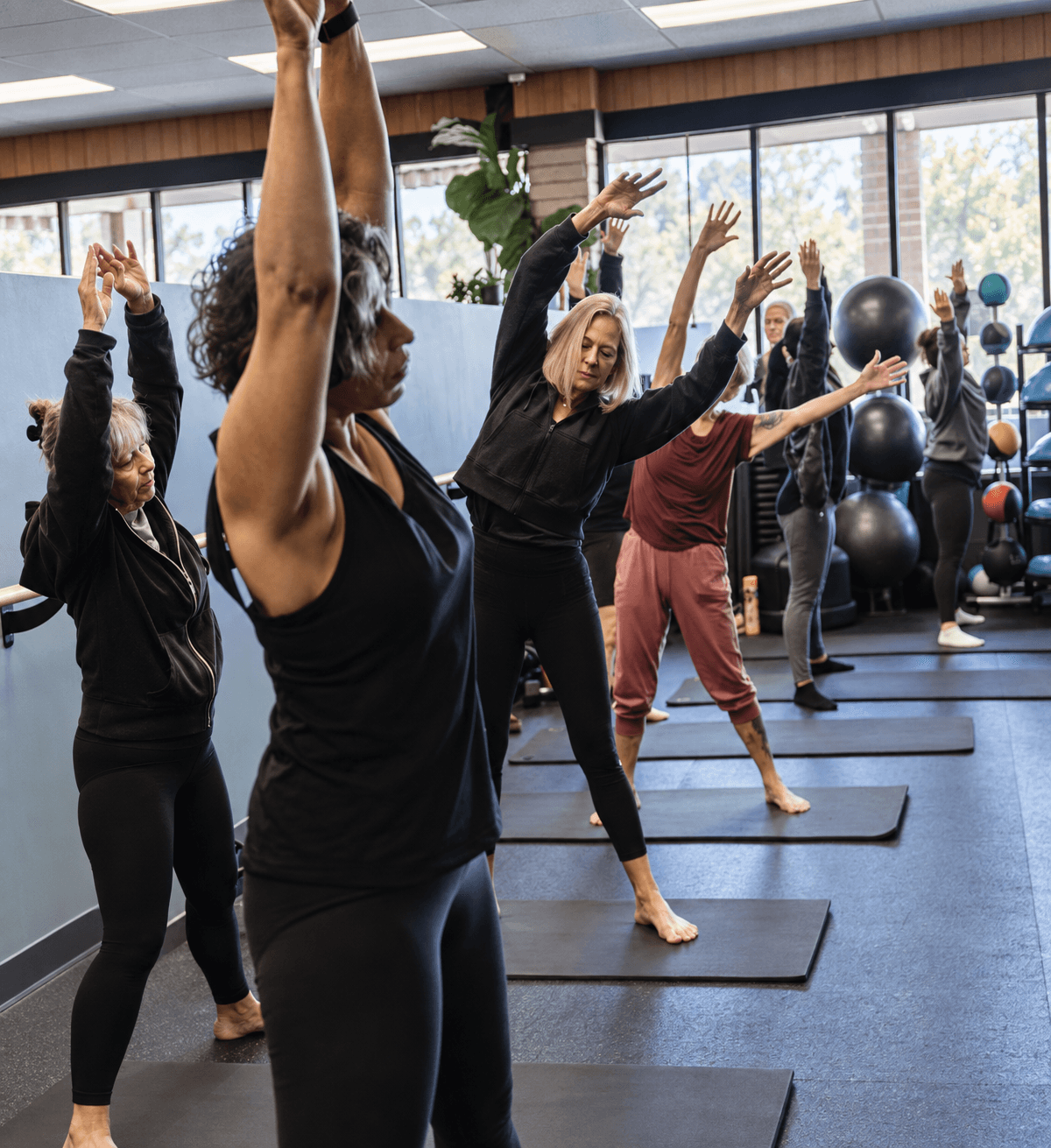 Group of people practicing yoga with arms raised in a fitness studio, large windows, and exercise equipment in background.