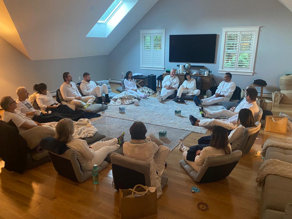 Group of people sitting in a circle in a room with a large TV, windows, and skylight, participating in a meeting or discussion.