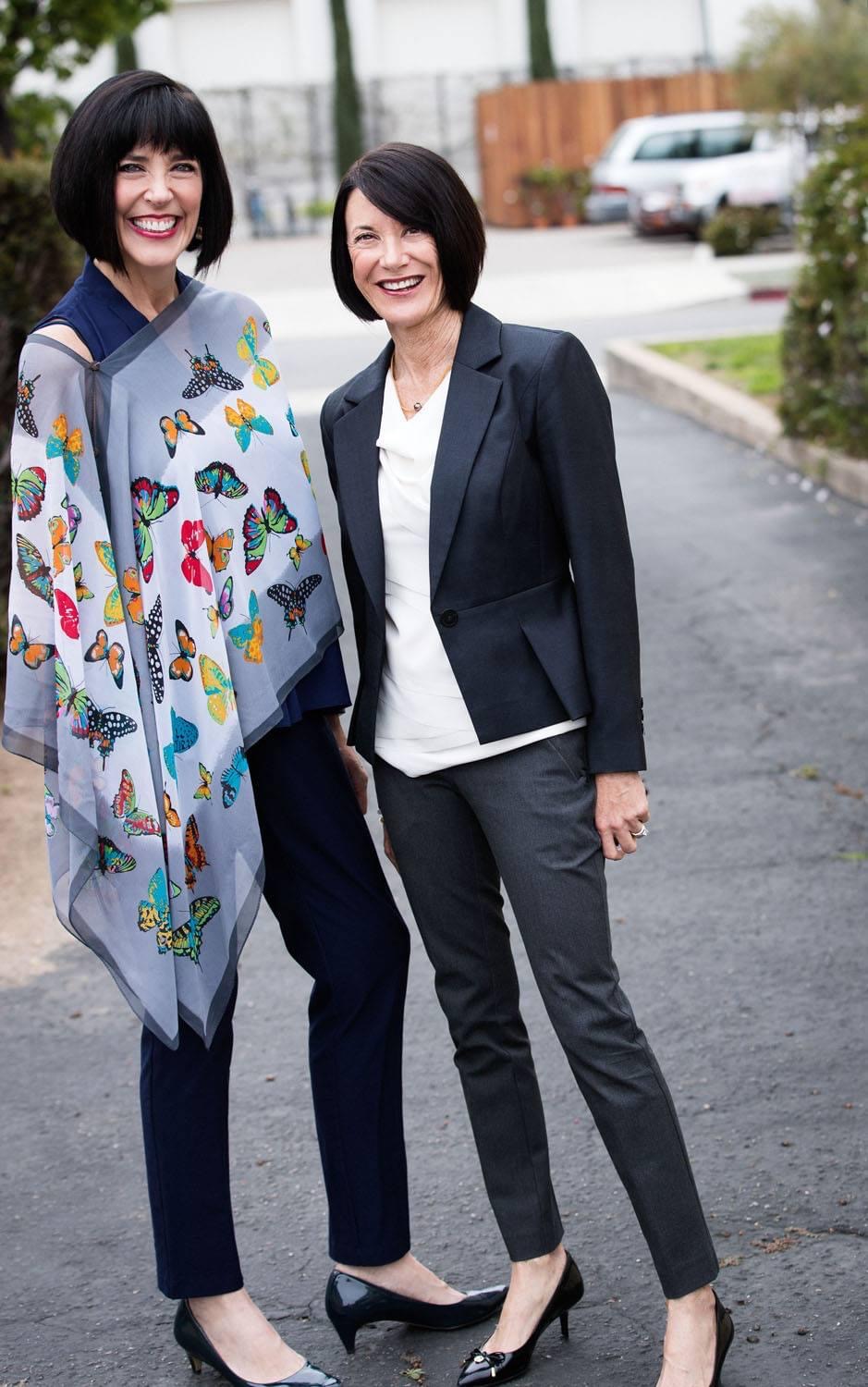 Two women standing outdoors, smiling, with a street and parked cars in the background.