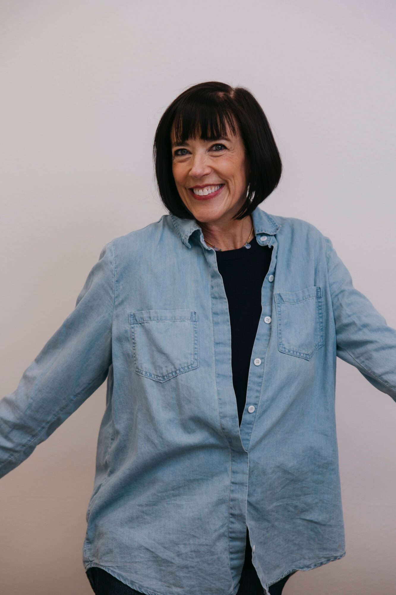 Smiling woman with short dark hair wearing a denim shirt and black top, standing against a plain background.