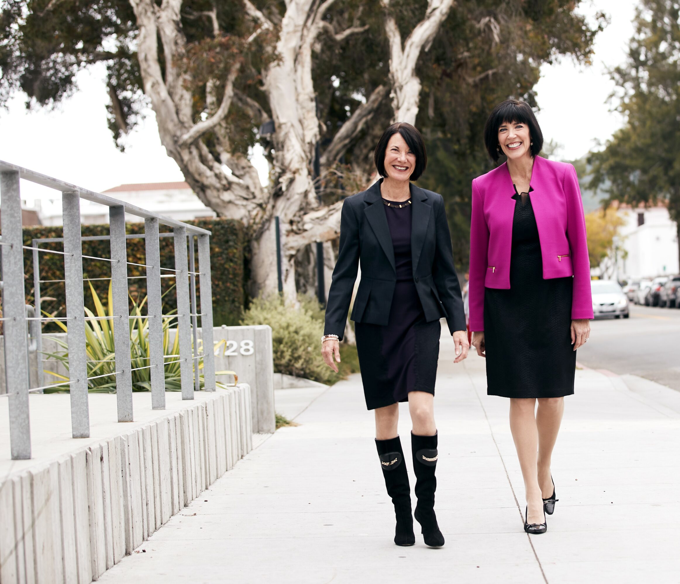 Two women walk on a sidewalk, smiling, with trees and a fence in the background.