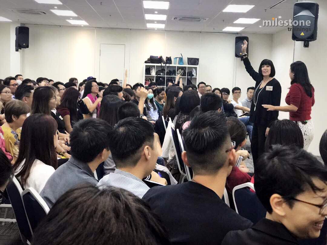 A large group of people seated in a room, listening to a speaker at the front, with a woman raising her hand.