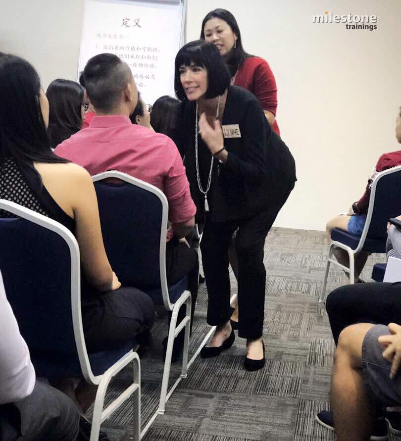 Woman with short black hair and black outfit crouches and speaks to seated group, with another woman standing behind her. Audience seated in chairs.