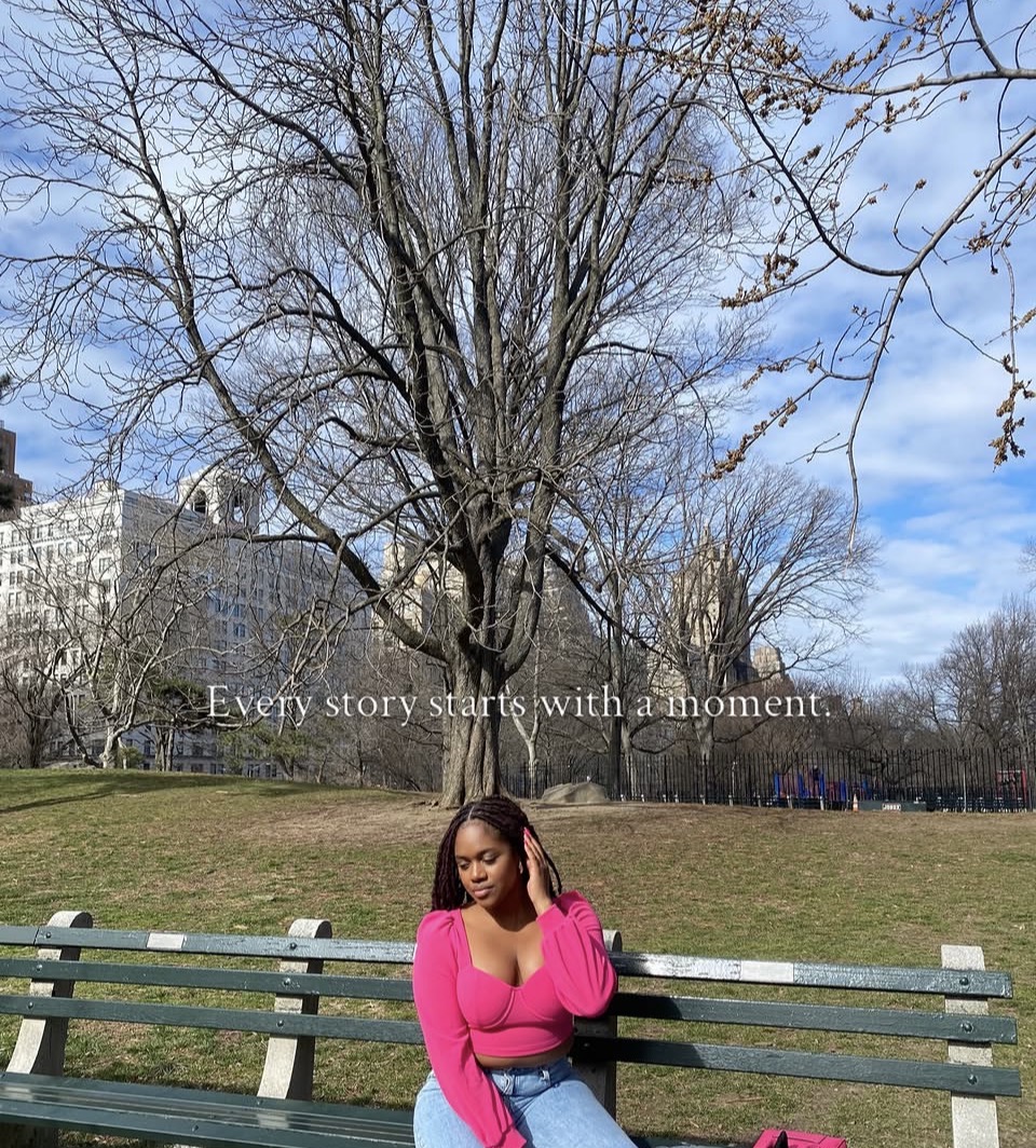 Woman in pink sweater sitting on a park bench with a large tree and blue sky in background.