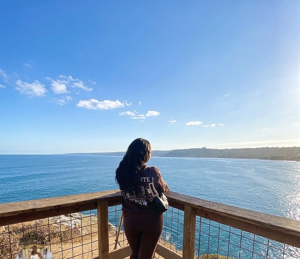 Woman with long dark hair standing on a wooden balcony overlooking the ocean under a clear blue sky.