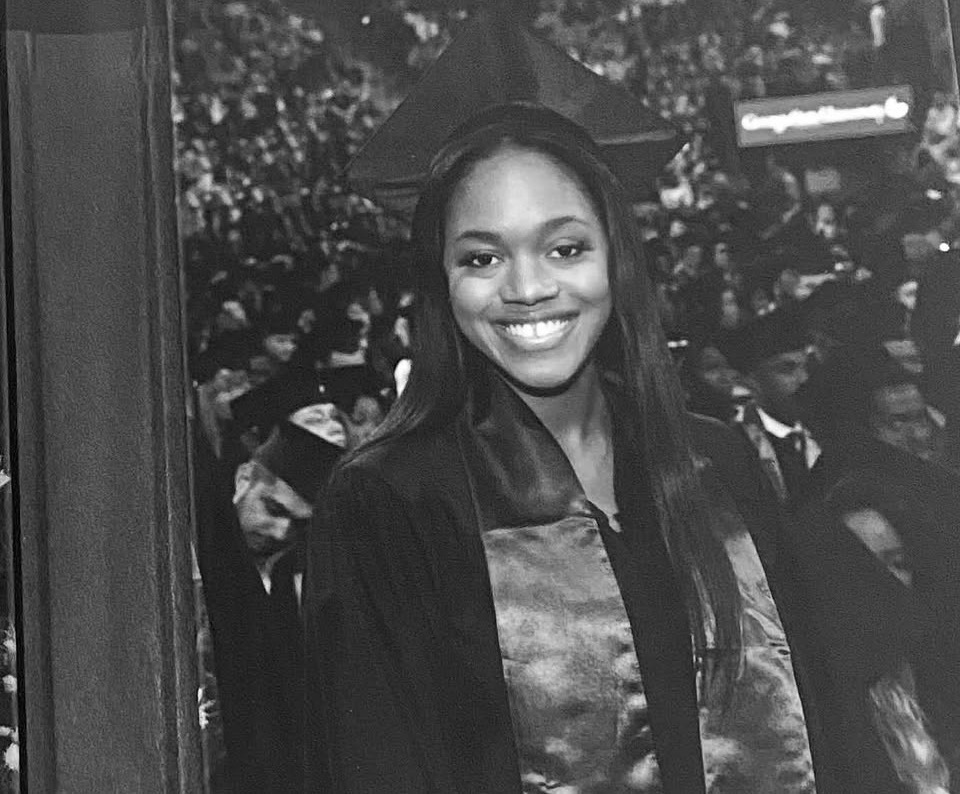 Young woman in graduation cap and gown smiling at ceremony, crowd in background, black and white photo.