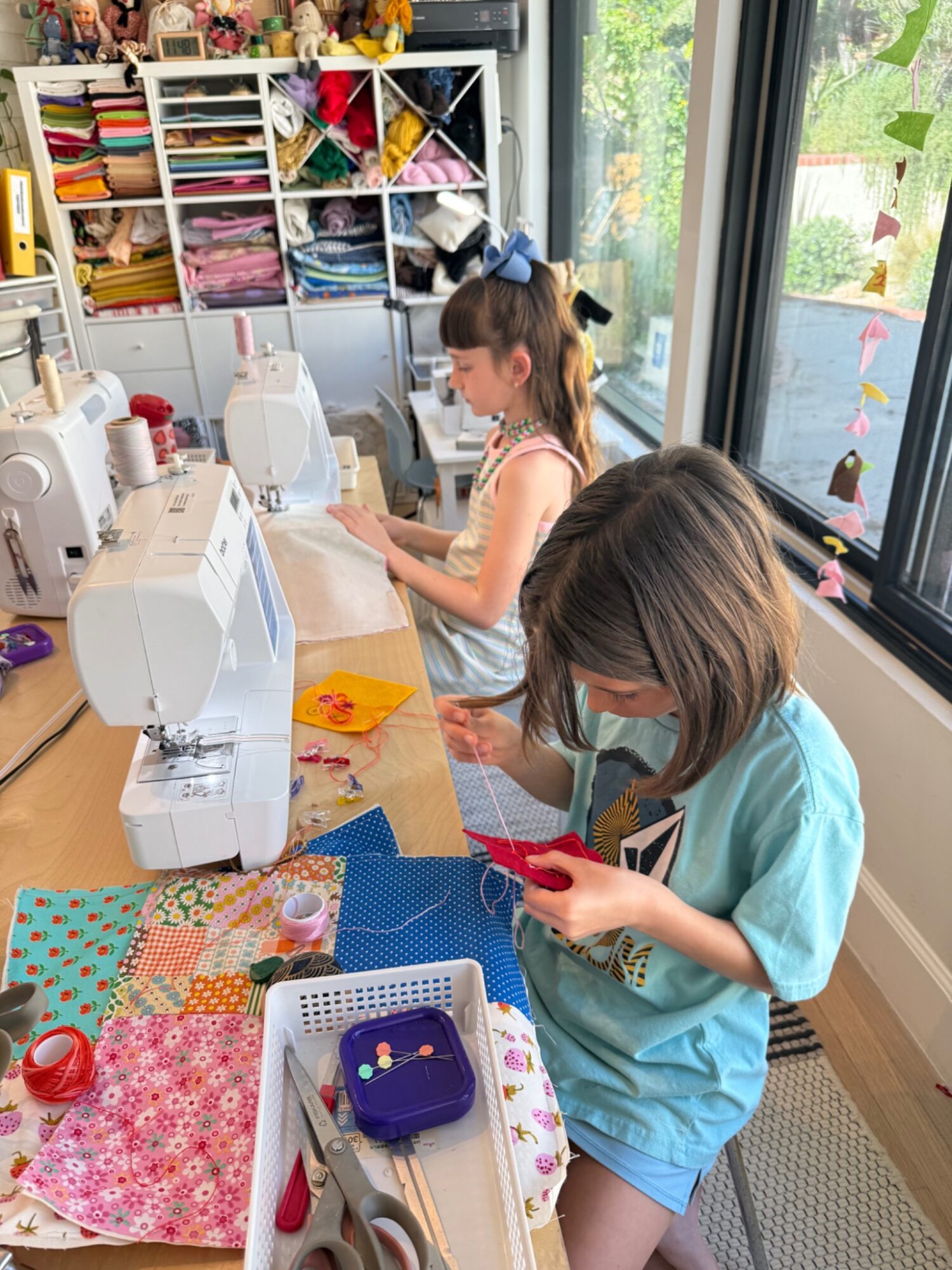 Two children sewing at a table with fabric, sewing machines, and sewing supplies, near large windows with plants outside.