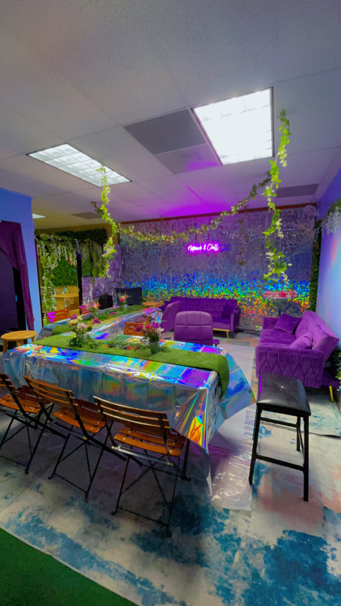 Indoor space with colorful furniture, hanging plants, and a vibrant wall, illuminated by ceiling lights and a neon sign.