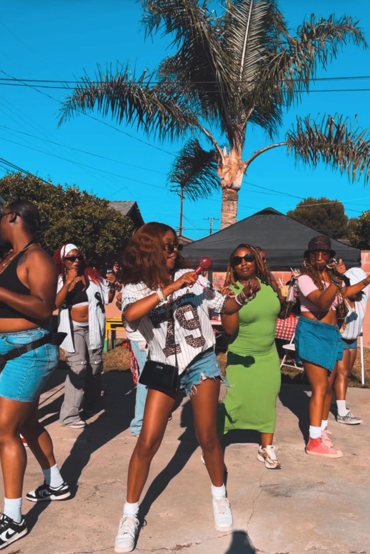 Group of people dancing outdoors with palm trees and tents in the background, sunny day, casual summer clothing.
