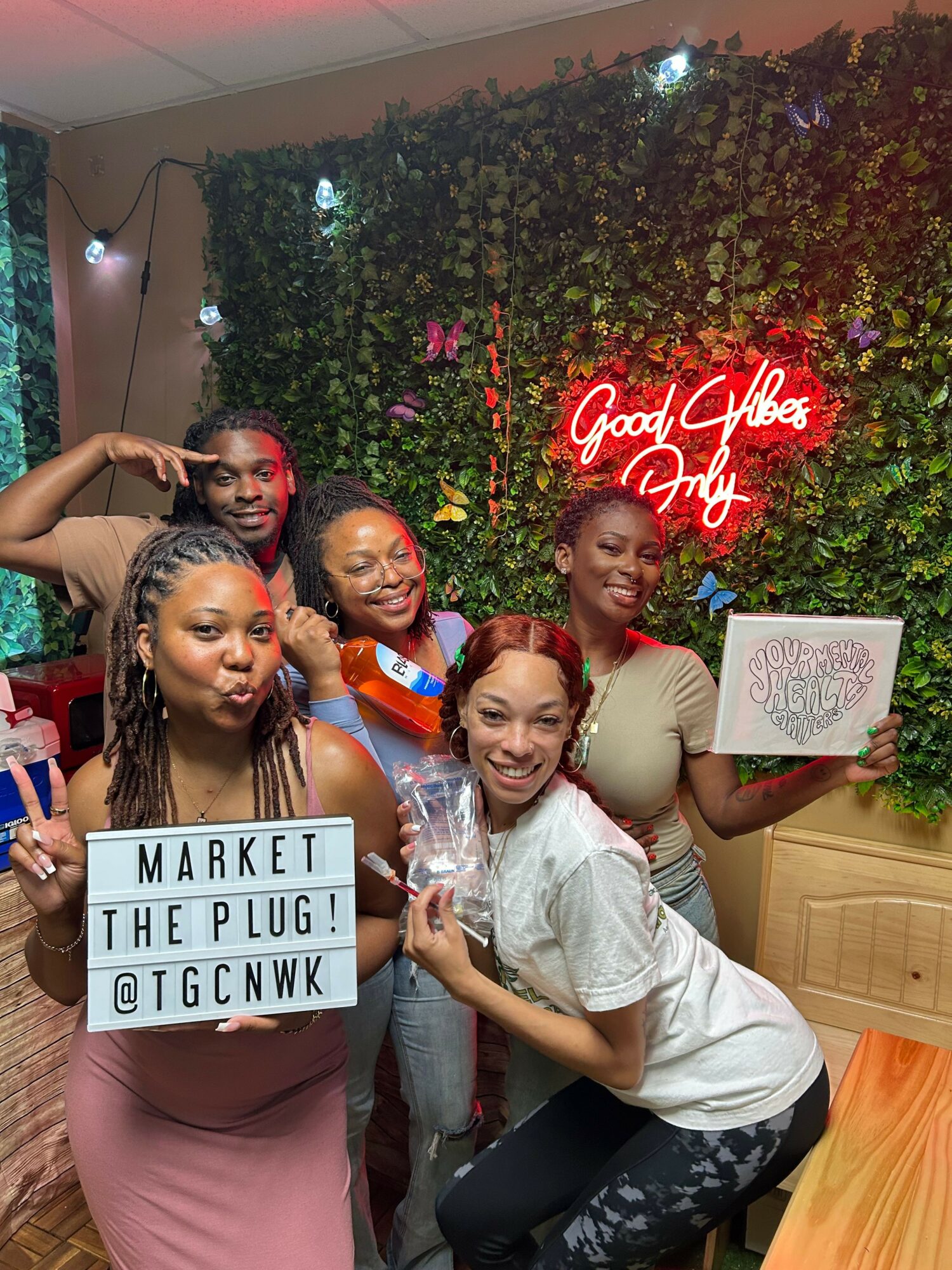 Four women celebrating outdoors at night, holding signs and drinks, with a neon sign and greenery in the background.