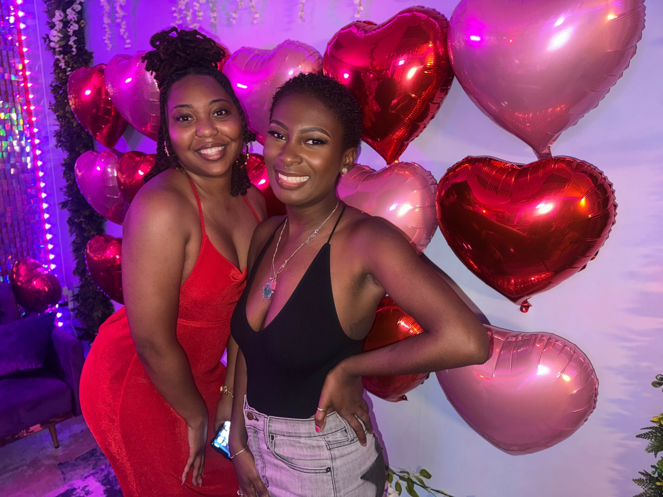 Two women smiling at a celebration with pink and red heart-shaped balloons in the background.