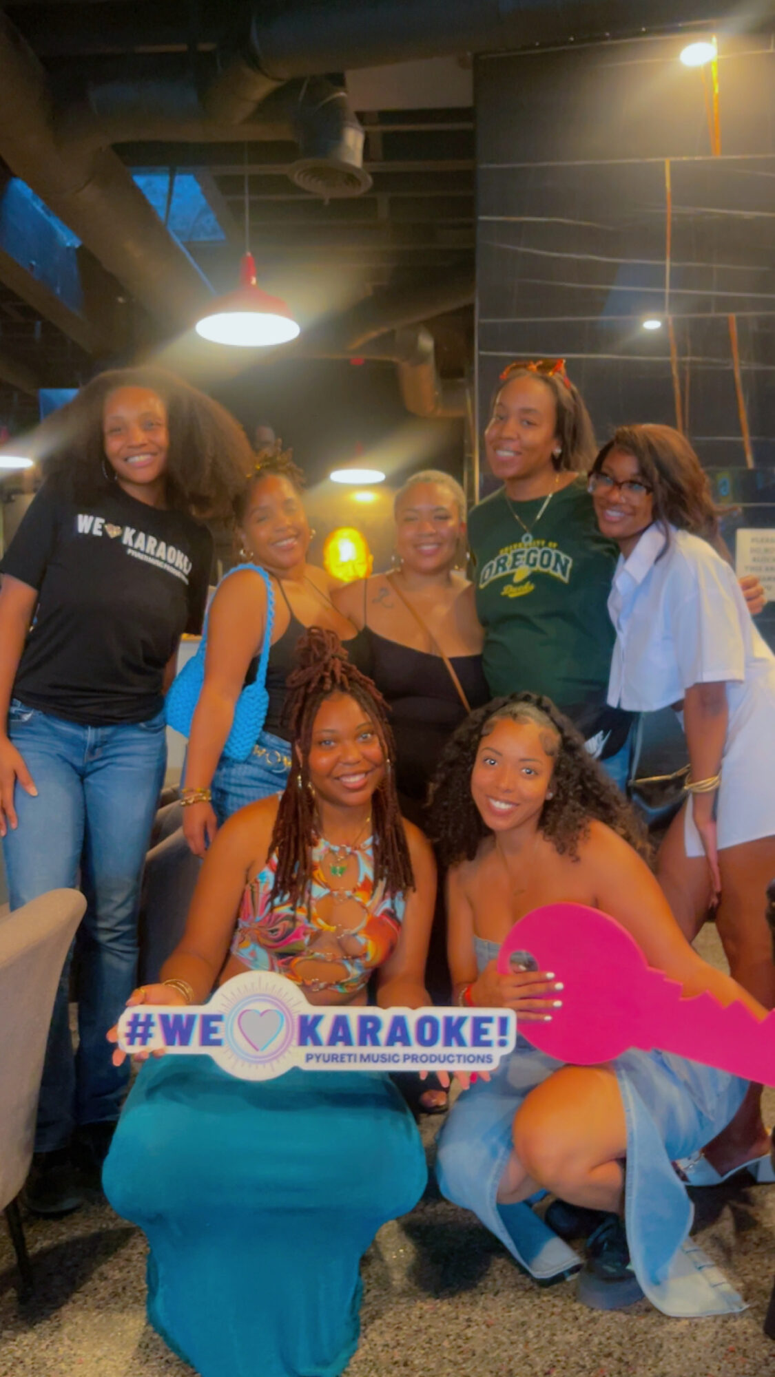 Group of six women smiling indoors, some sitting and some standing, with a karaoke sign, in a dimly lit setting.