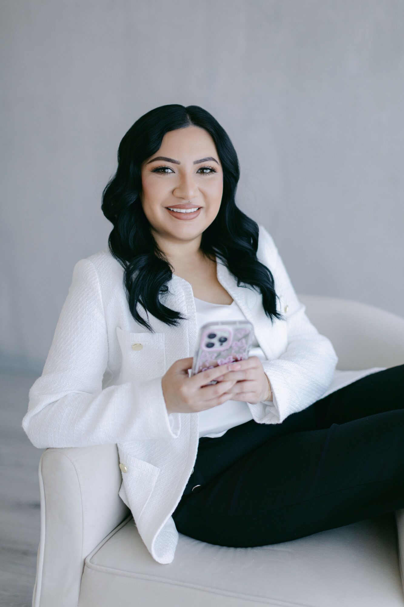 Young woman with dark wavy hair smiling, holding a phone, sitting on a white couch against a plain background.