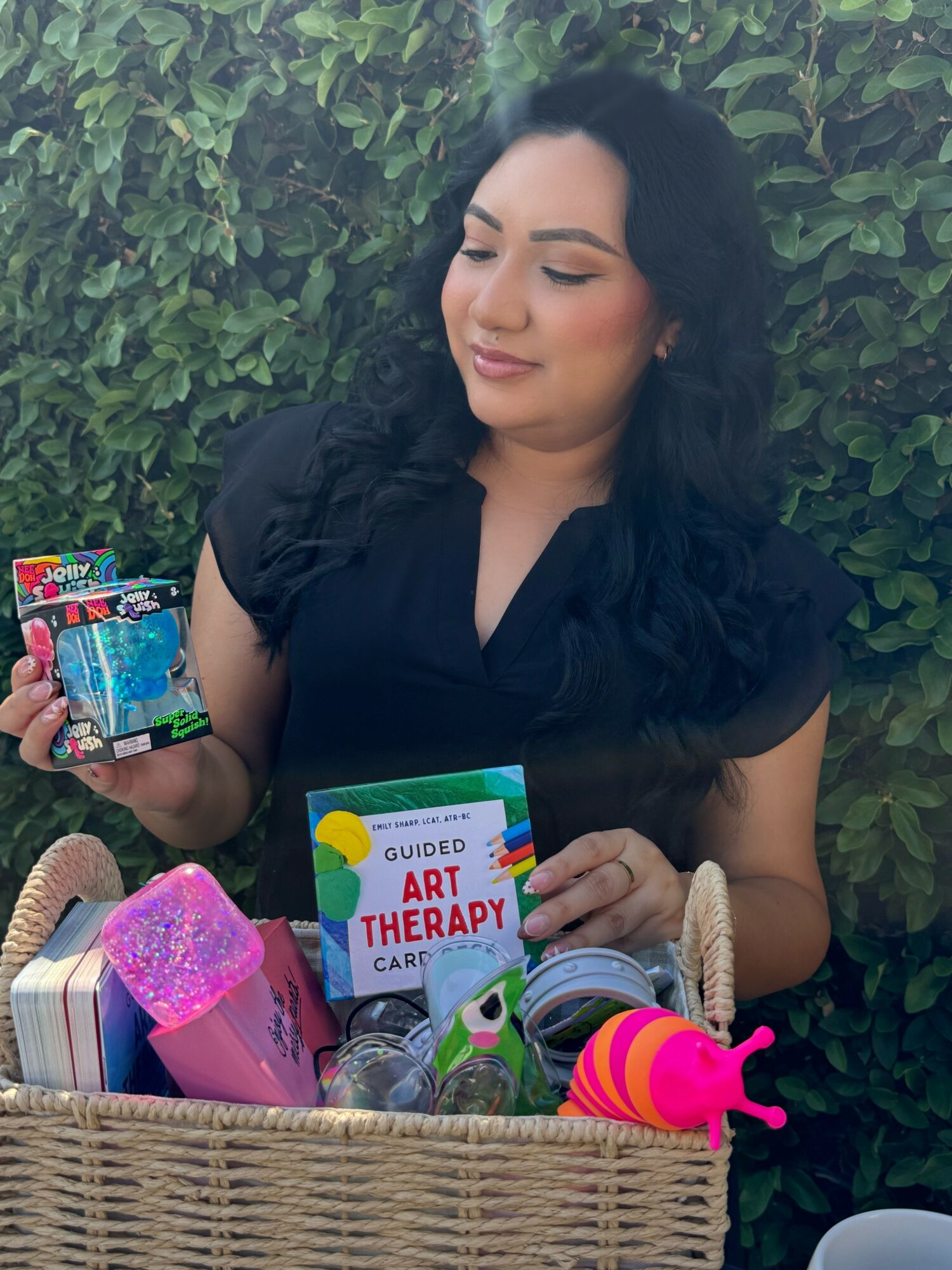 Woman with dark hair holding art therapy cards, surrounded by art supplies in a basket, outdoors with green foliage.