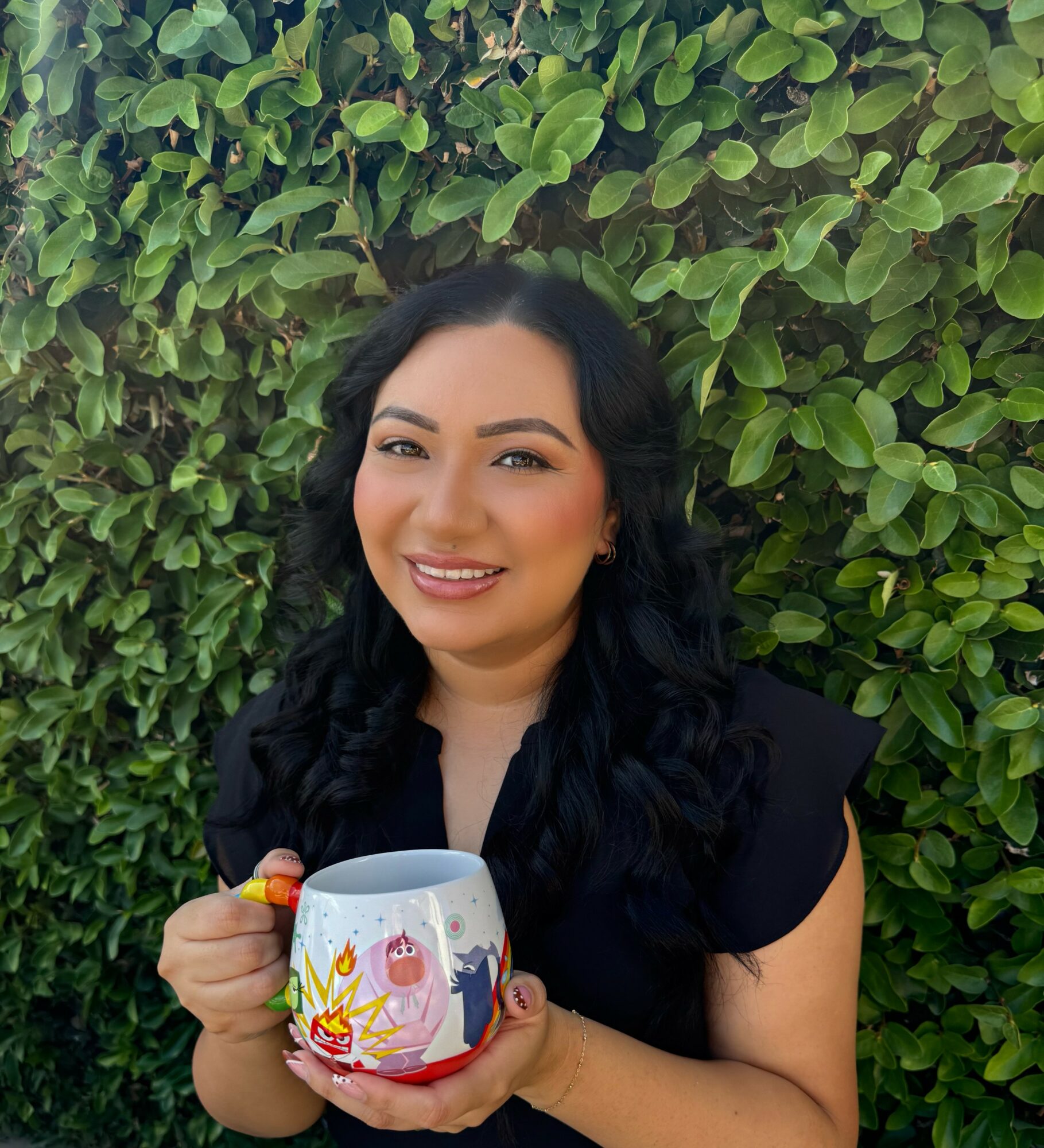Woman with dark wavy hair smiling, holding a colorful mug, standing in front of green leafy background.
