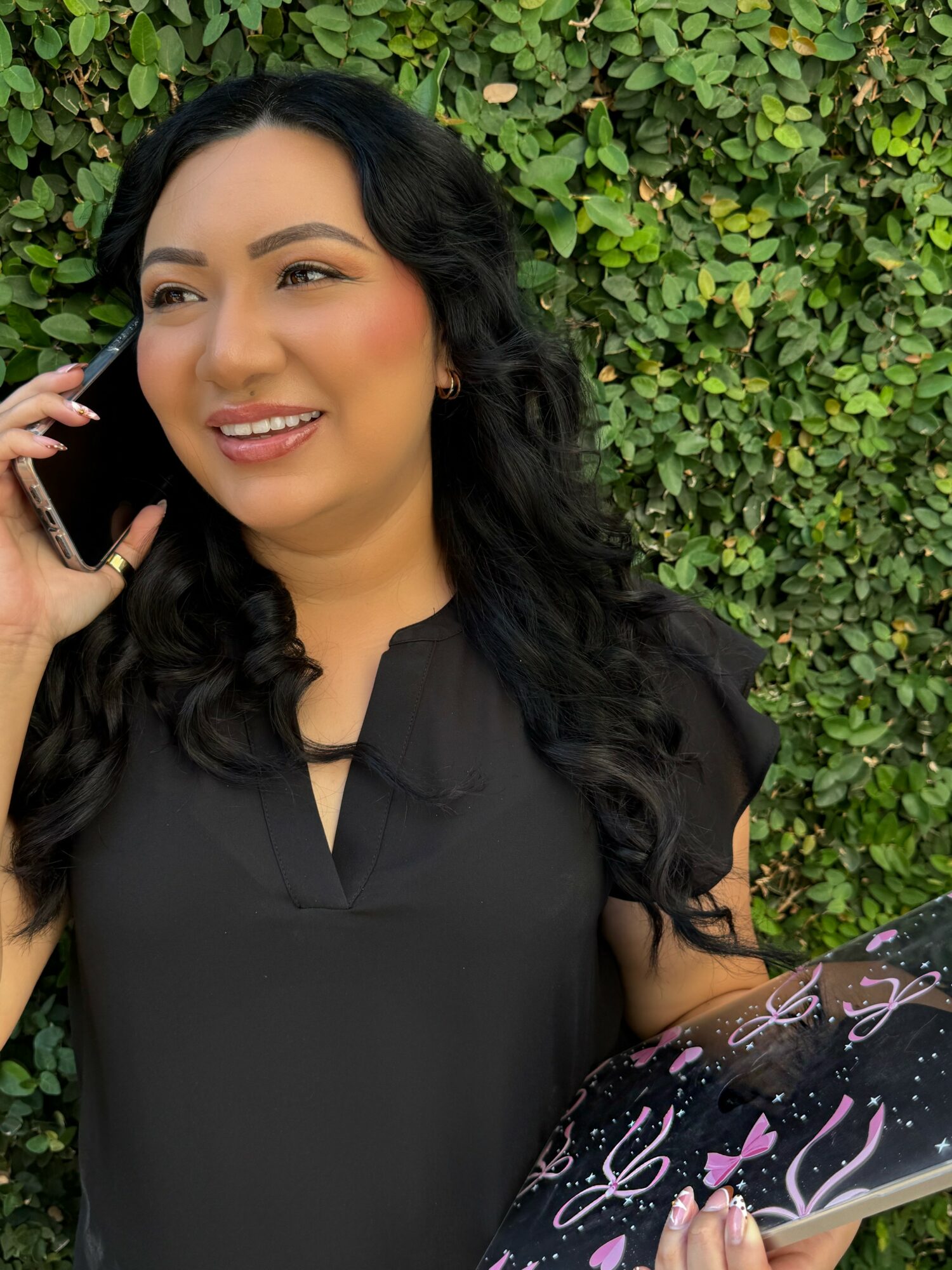 Woman with long dark hair smiling, talking on phone, standing in front of green leafy background.