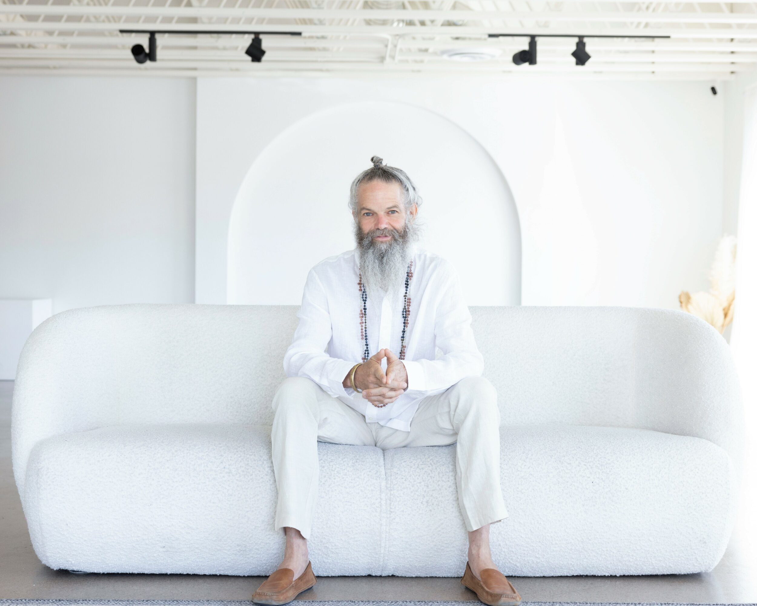 Man with a beard and long hair sitting on a white sofa in a bright, minimalist room with white walls and ceiling.