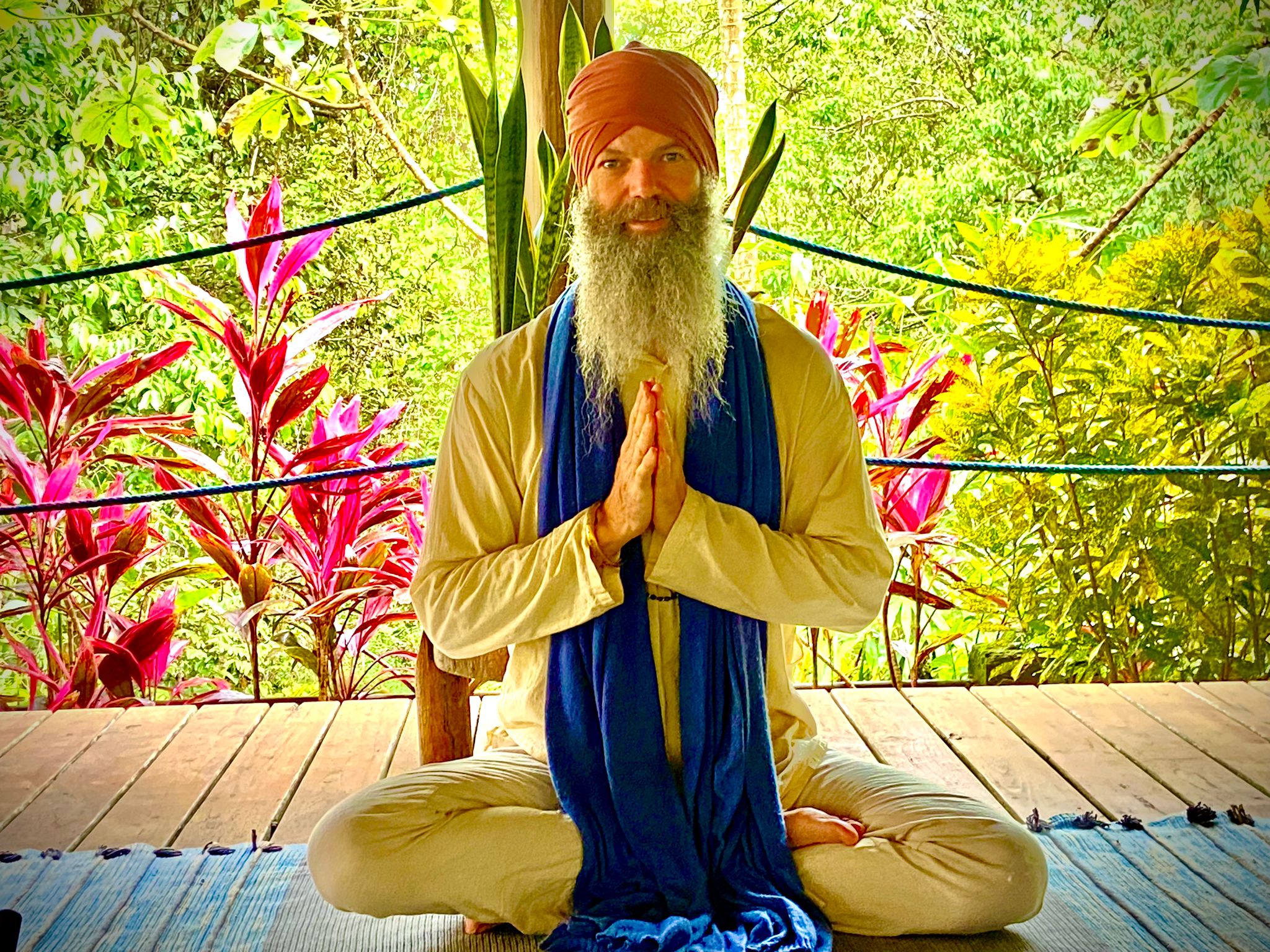 A man with a beard and turban sits cross-legged on a wooden platform, hands in prayer position, surrounded by greenery and pink plants.