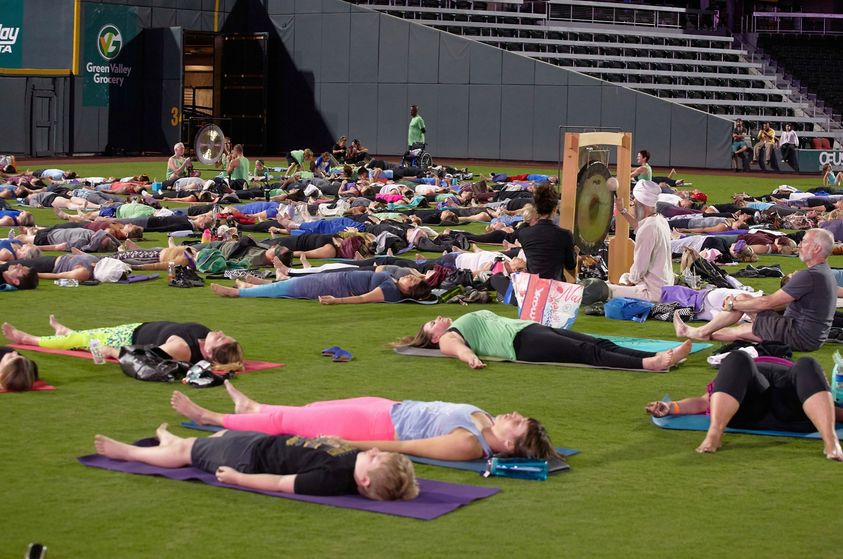 People practicing yoga on a field with mats, lying down in a relaxed pose, with some standing and sitting nearby.