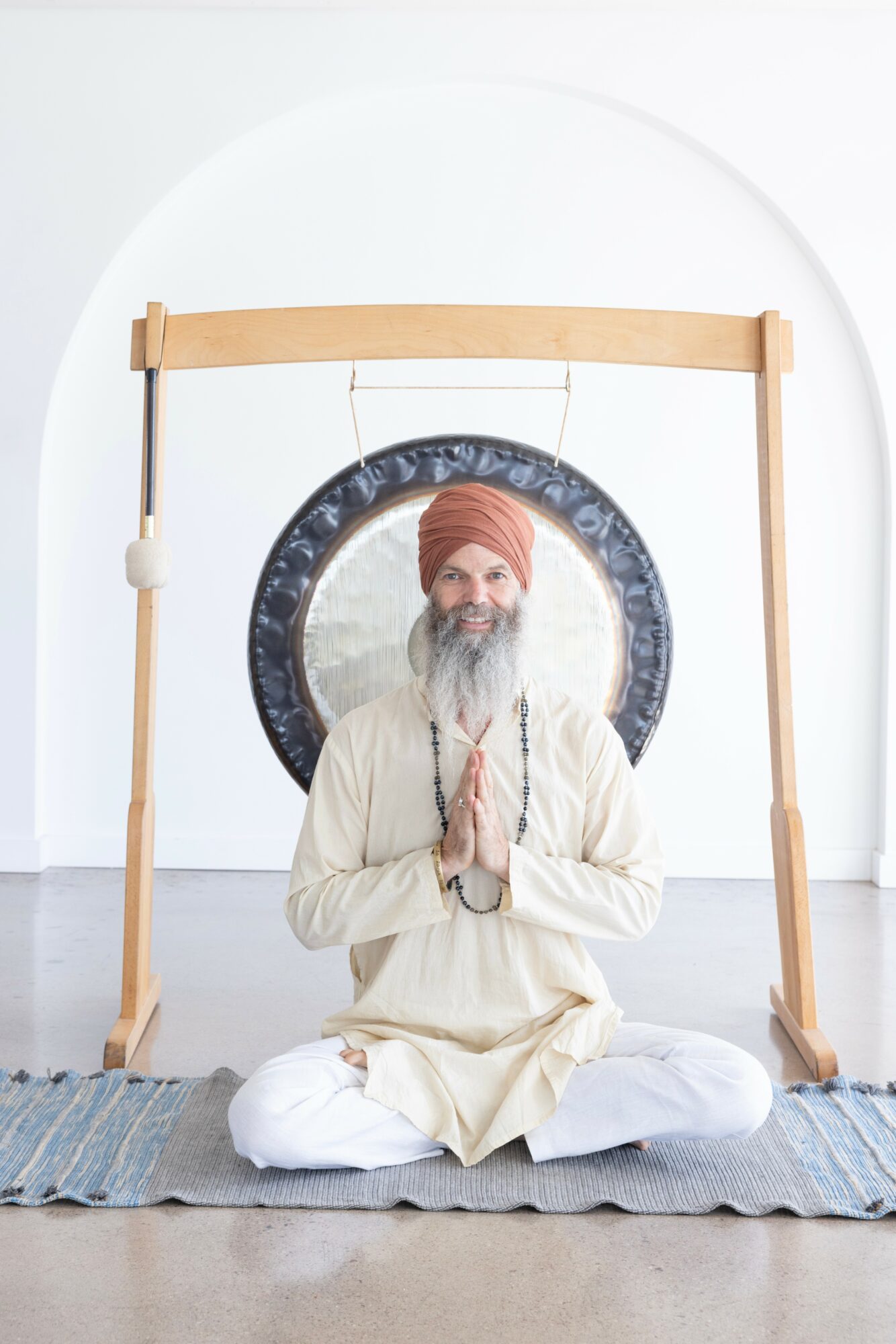 Person in traditional attire sitting cross-legged with hands in prayer position, in front of a large gong, indoors.