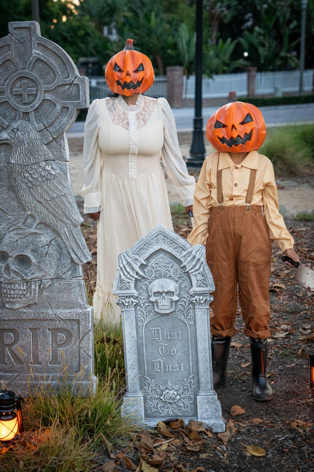 Person and child with pumpkin masks standing between Halloween tombstones outdoors, trees and a fence in background.