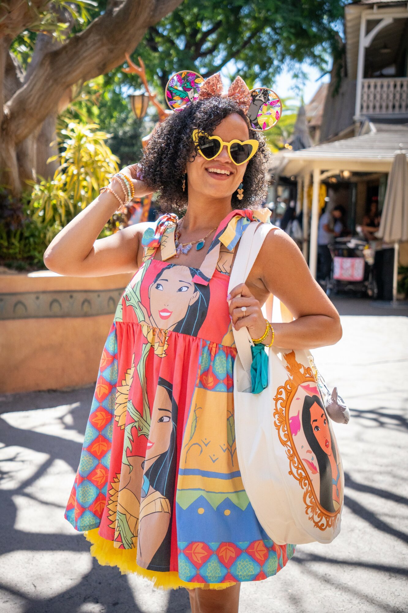 Woman wearing colorful dress with cartoon faces, yellow heart-shaped sunglasses, and floral headband, smiling outdoors.