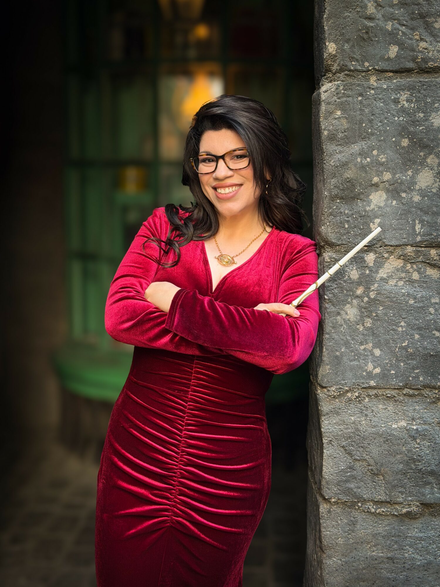 Woman in red dress with glasses smiling, holding a pointer, standing next to a stone wall.