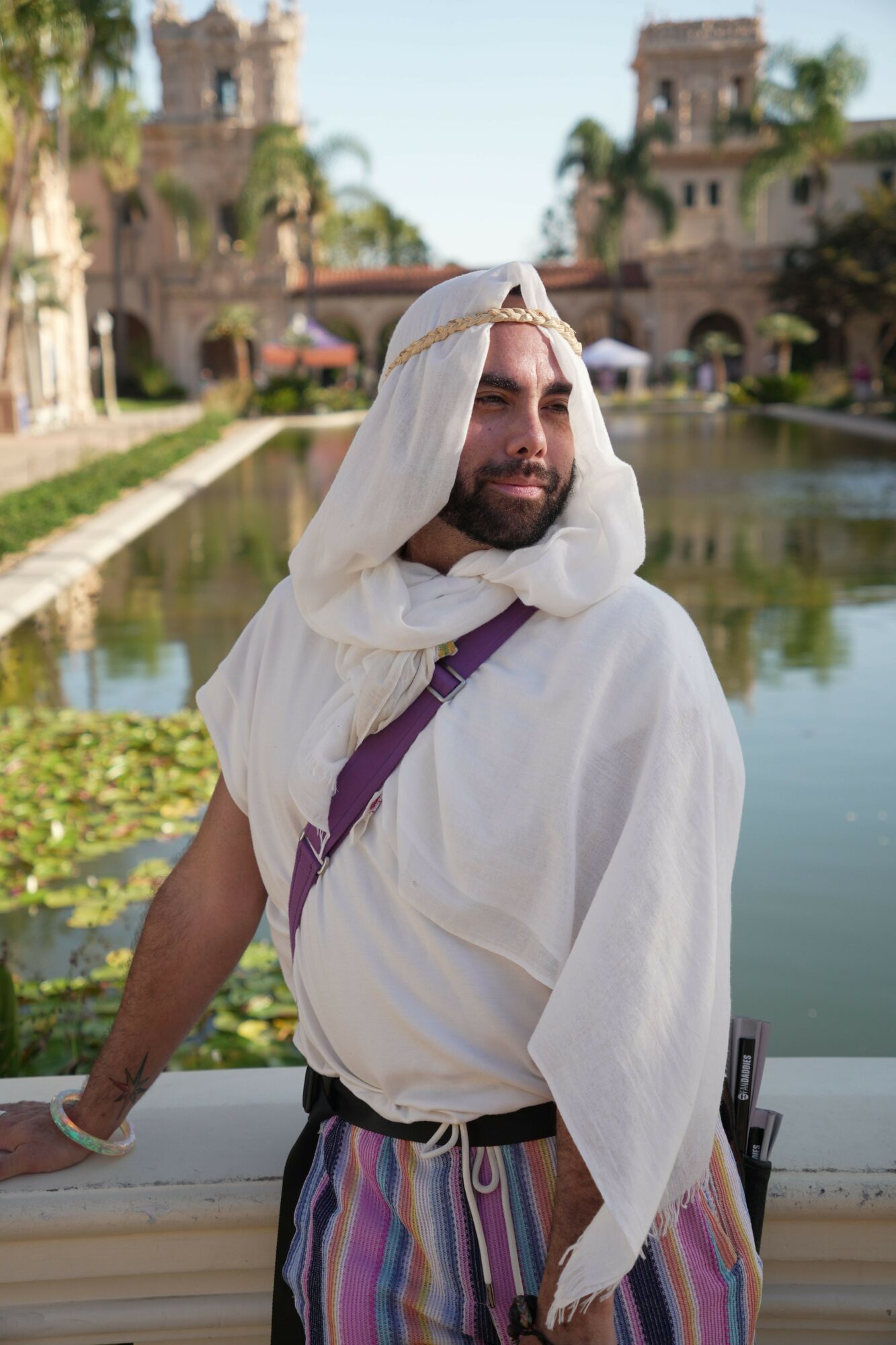 Person wearing a white head covering and glasses, standing outdoors near water with buildings in background.