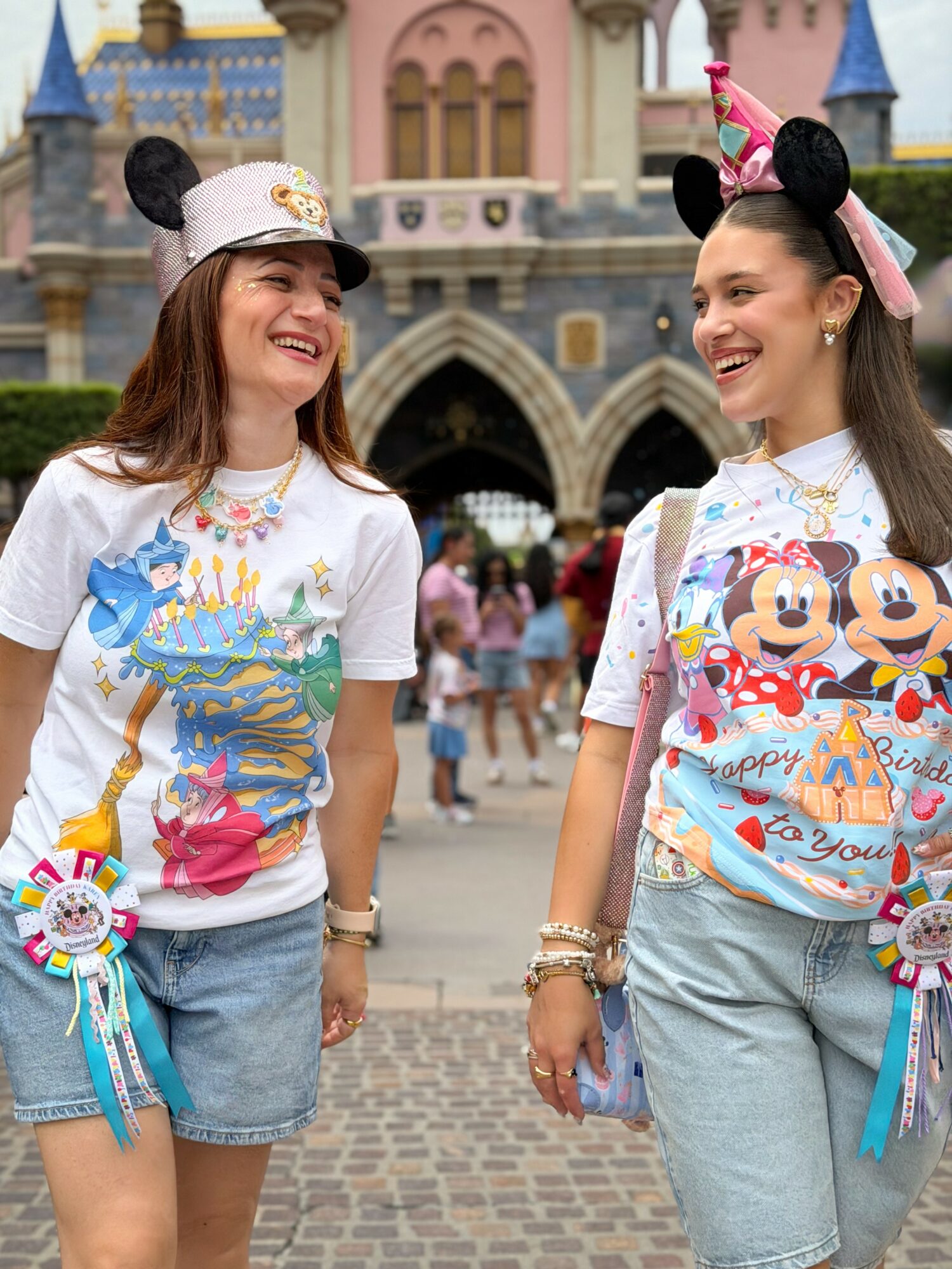 Two women wearing Mickey Mouse ears hats and colorful Disney-themed shirts, smiling at each other in front of a castle.