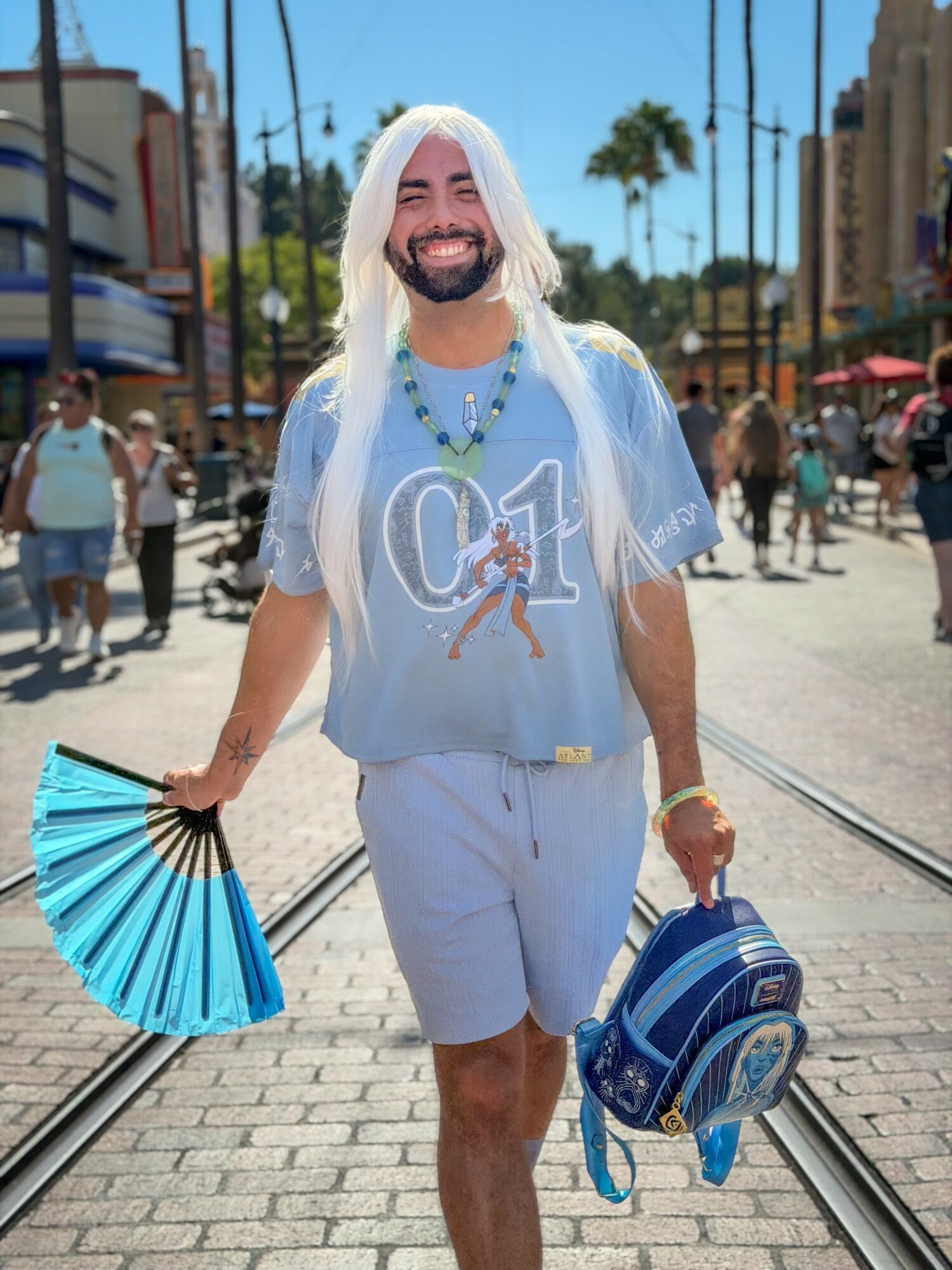 Person with long white hair smiling, wearing light blue outfit, holding a blue fan and backpack, walking outdoors.