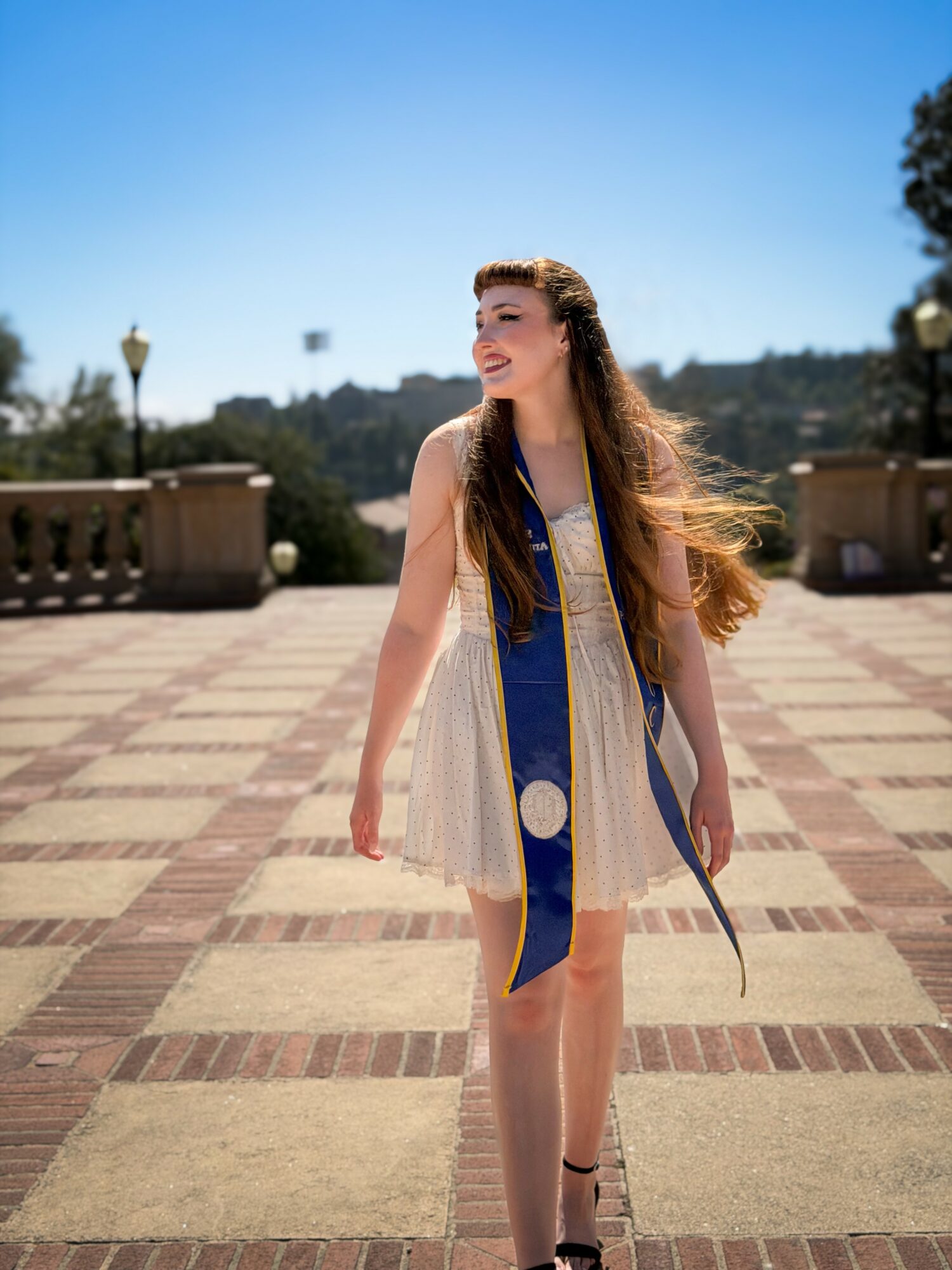 Young woman with long hair walking outdoors on a sunny day, smiling, wearing a white dress and a blue sash.
