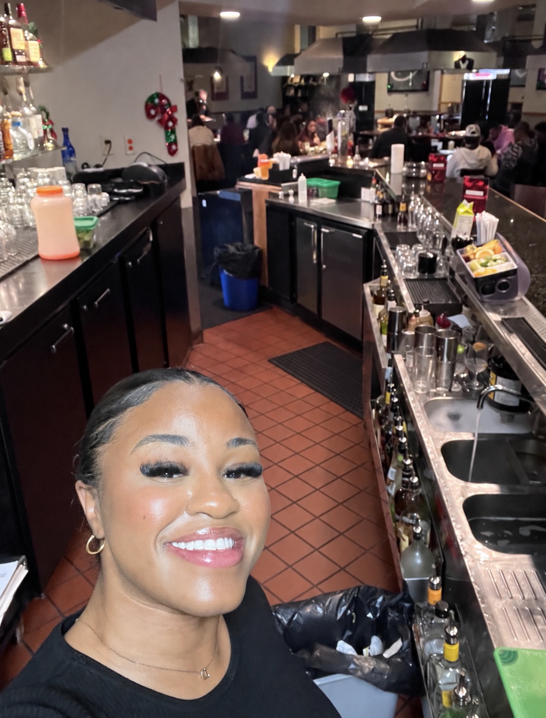Smiling woman taking a selfie in a bar or restaurant with a busy counter and patrons in the background.