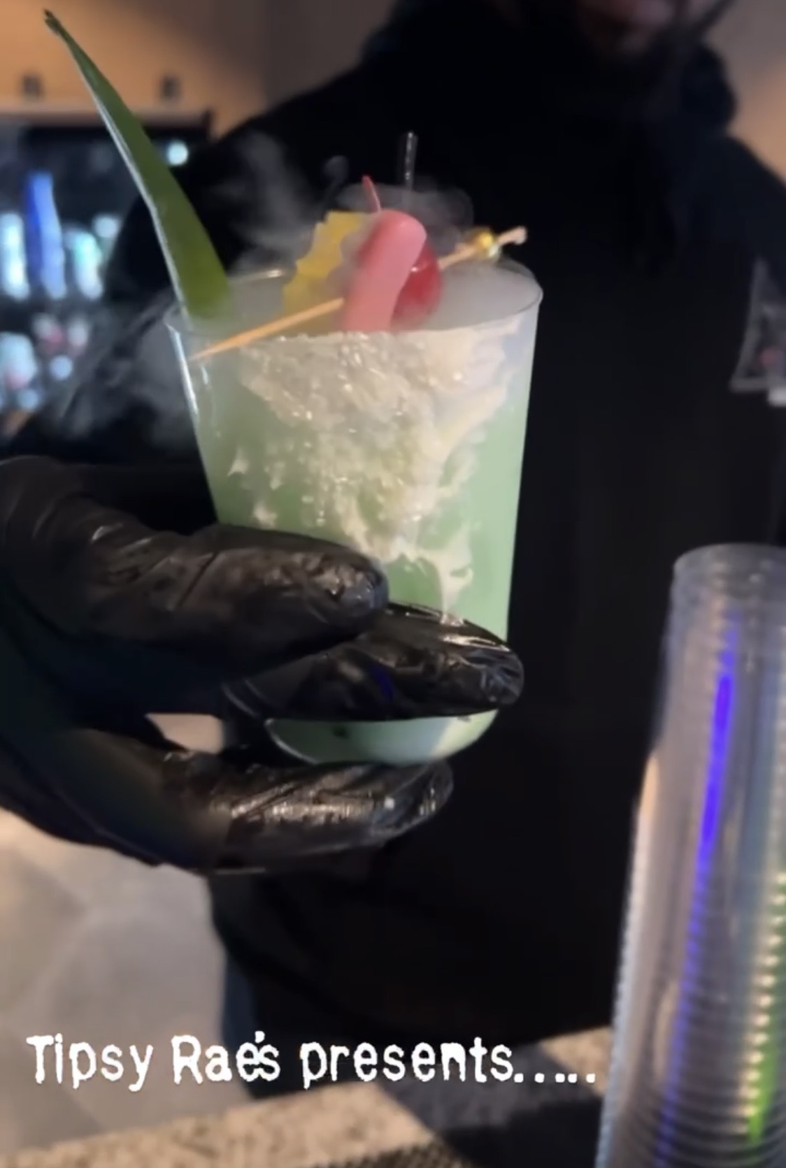 Person wearing black gloves holding a colorful cocktail with fruit garnishes, background is dark and blurry.