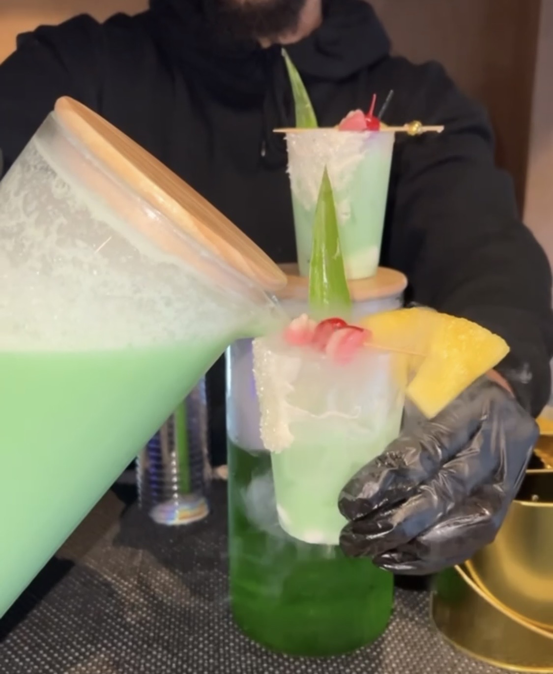 Person wearing black gloves holds a tall glass with a green layered drink, garnished with fruit and a straw.