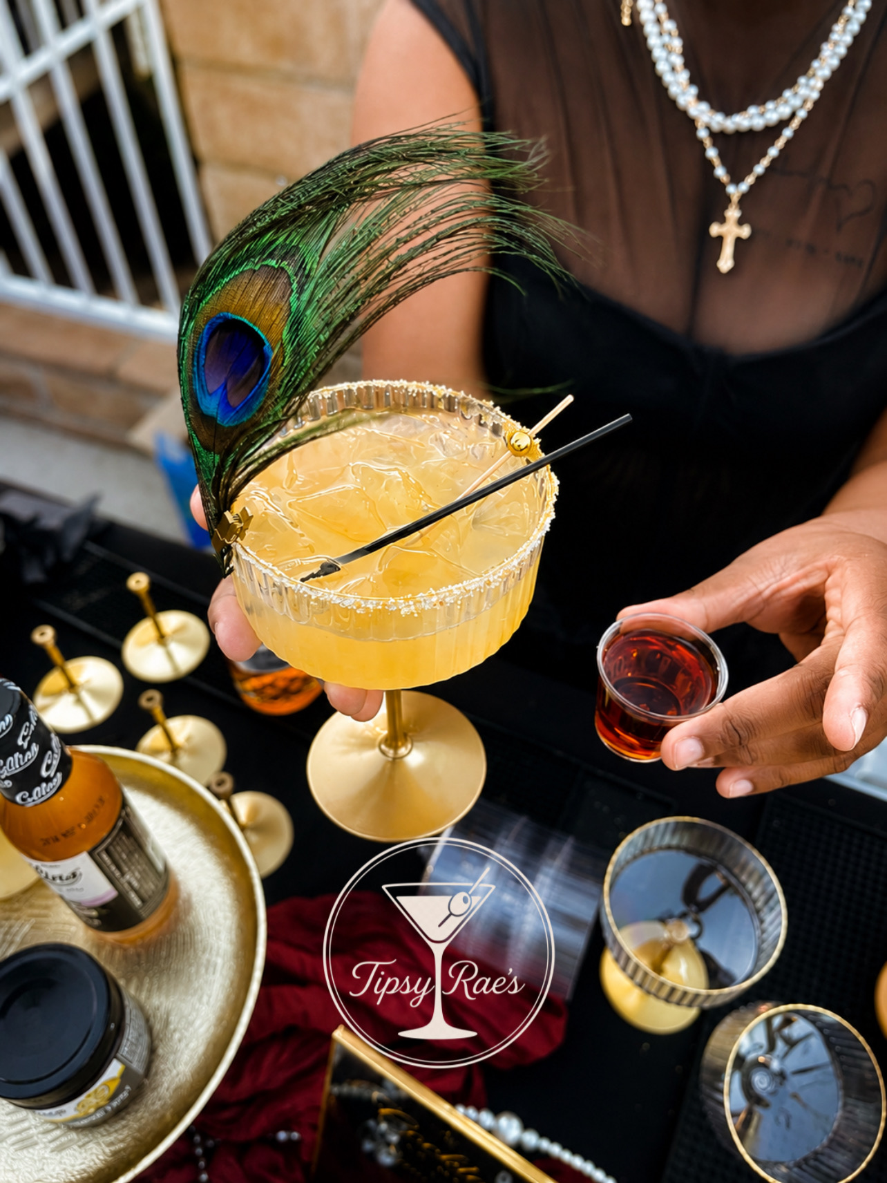 Person holding a cocktail with a peacock feather garnish, wearing a cross necklace, with drinks and bar items nearby.