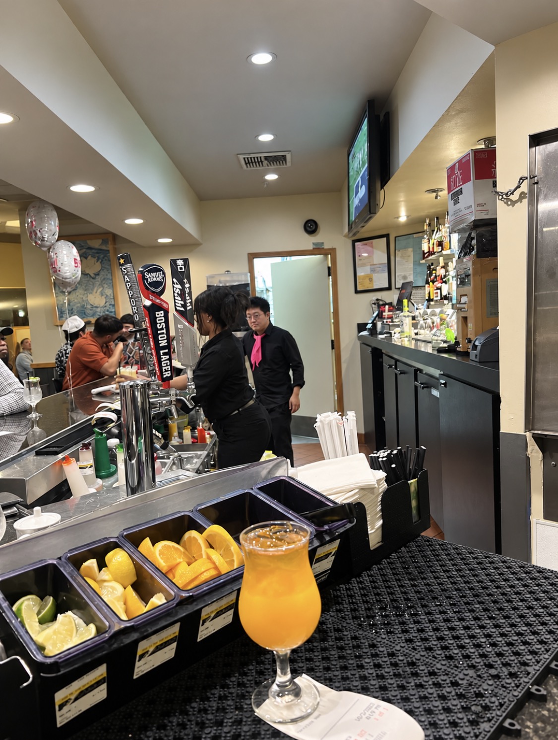 Bar counter with a glass of orange drink, fruit slices, and bar equipment, with people in the background inside a cafe or bar.