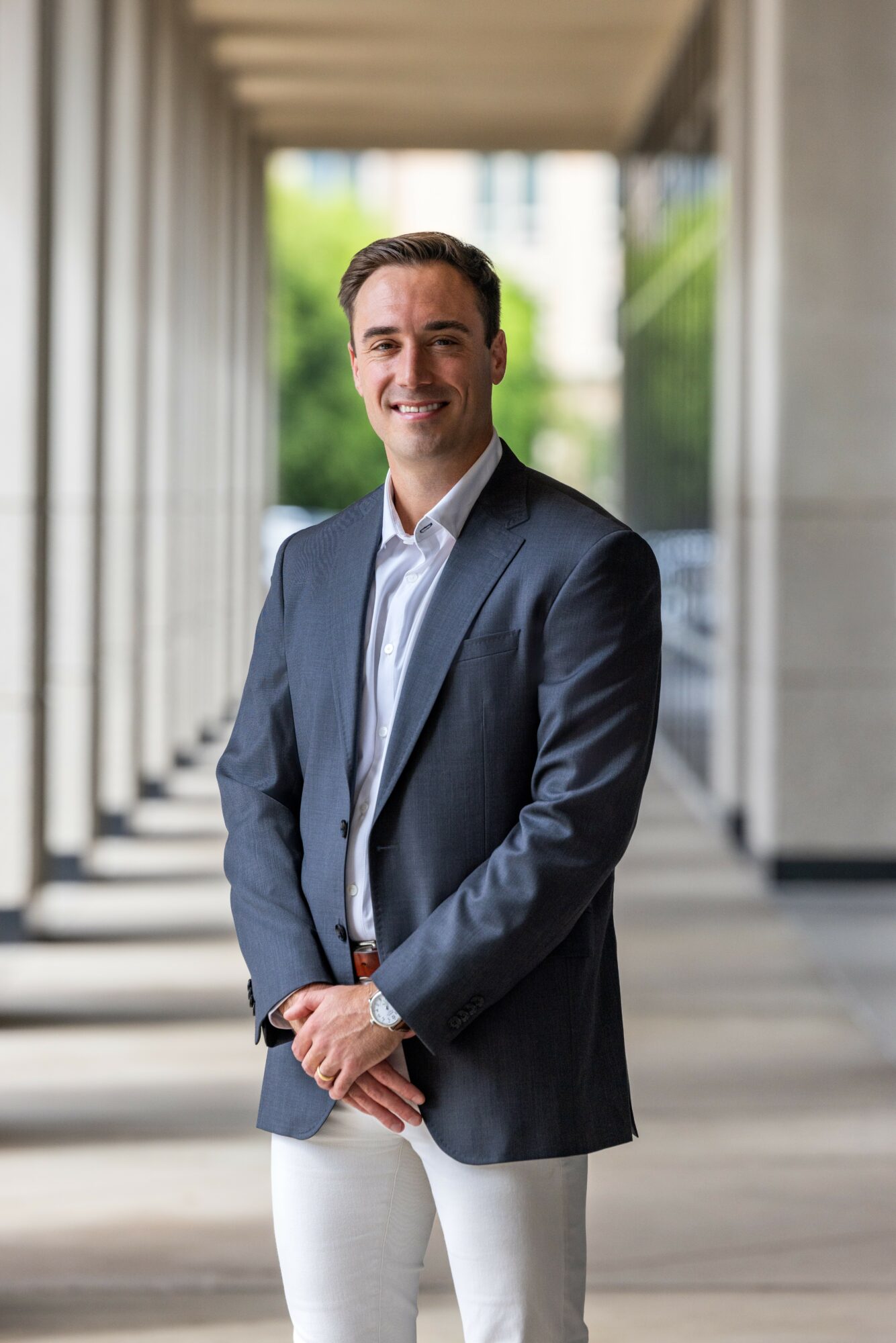 Man in a suit standing outdoors with columns and greenery in background.