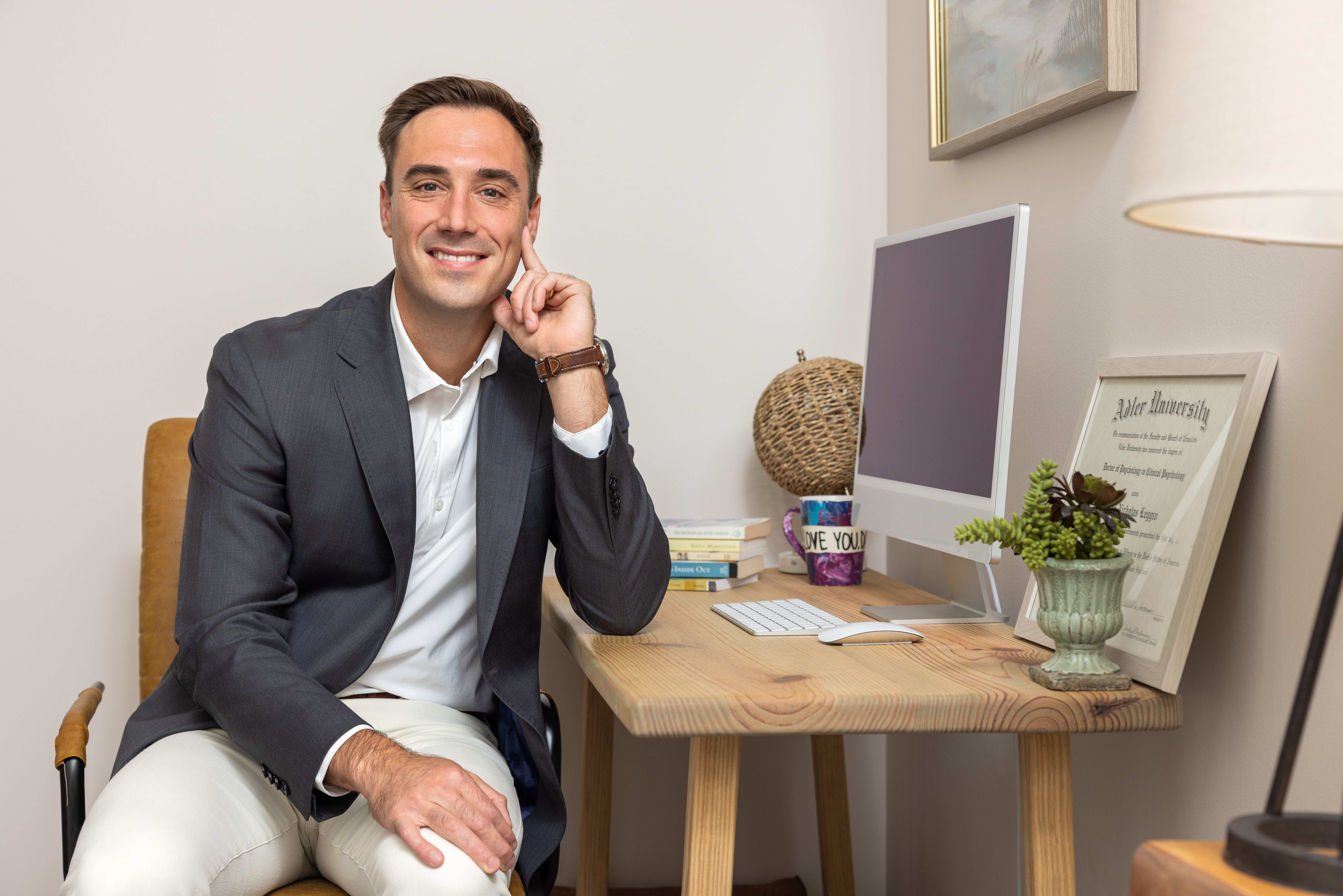 Man in a suit sitting at a desk with a computer, smiling, in a well-lit room.