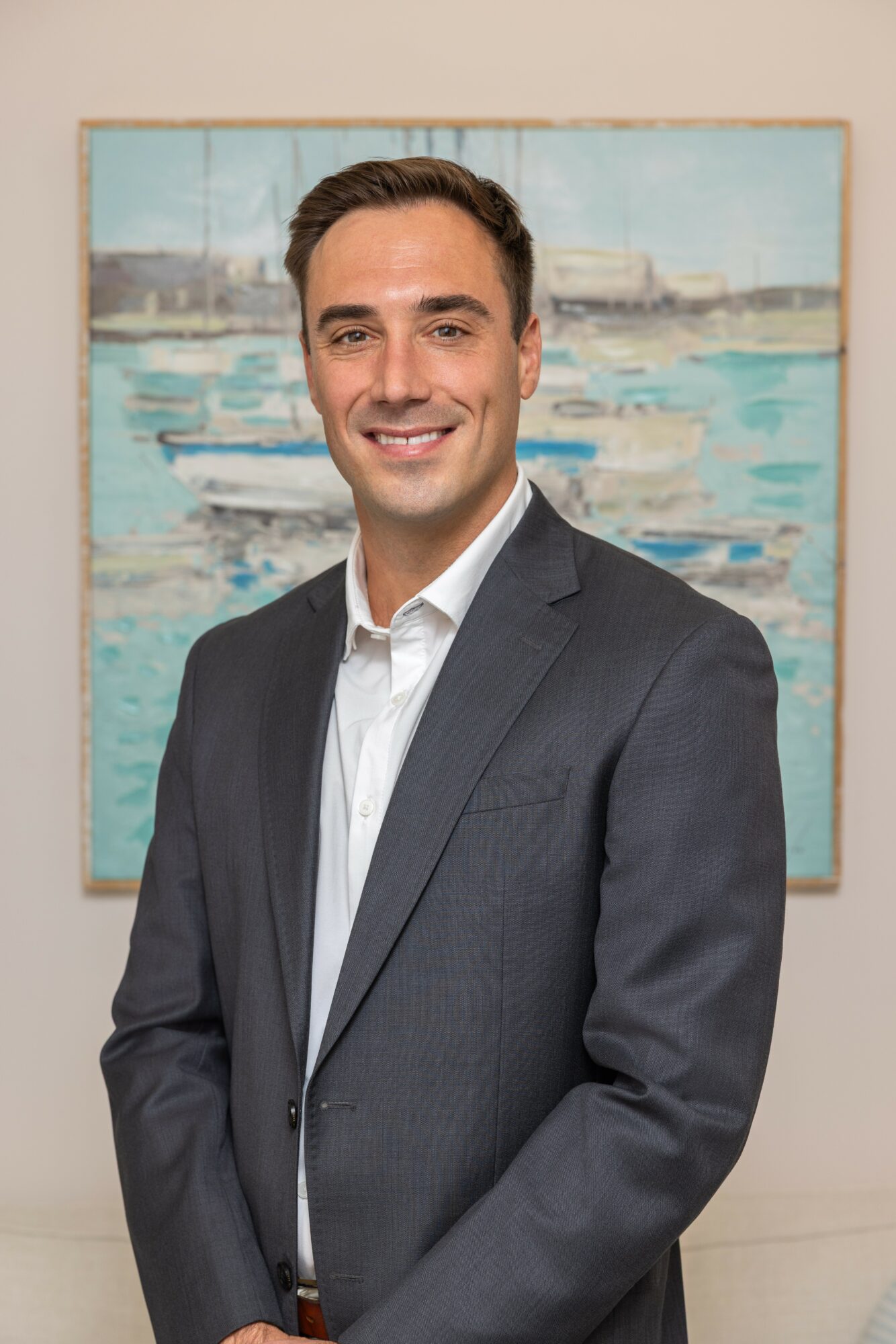Man in a gray suit and white shirt smiling, standing in front of a painting of boats and water.