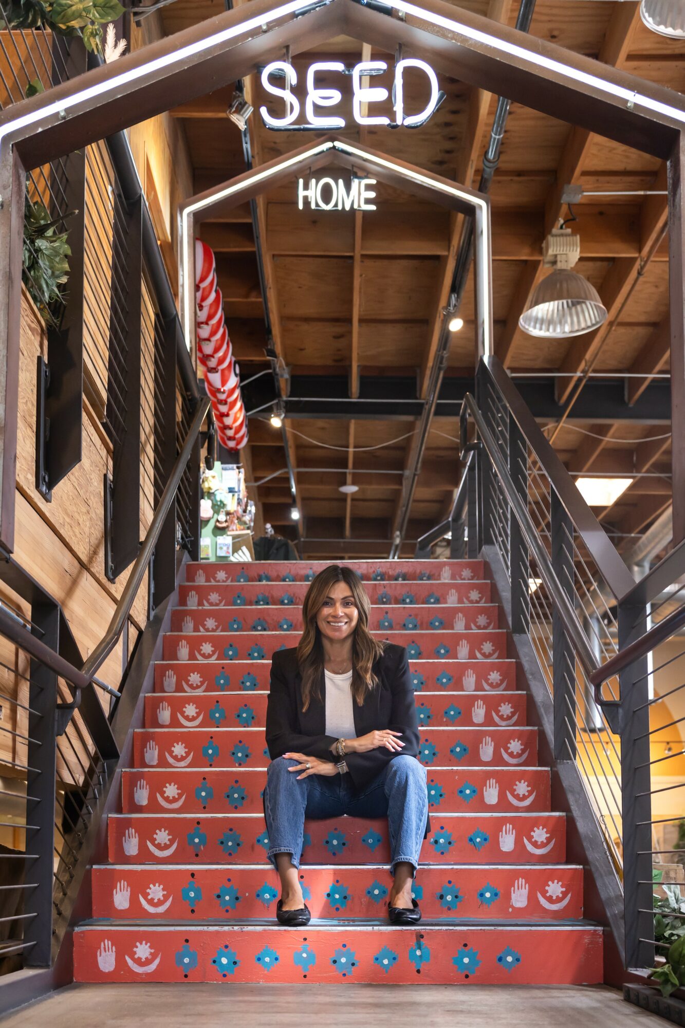 Woman sitting on red staircase with blue and white patterns inside a wooden building, with signs reading 'SEED' and 'HOME' above.